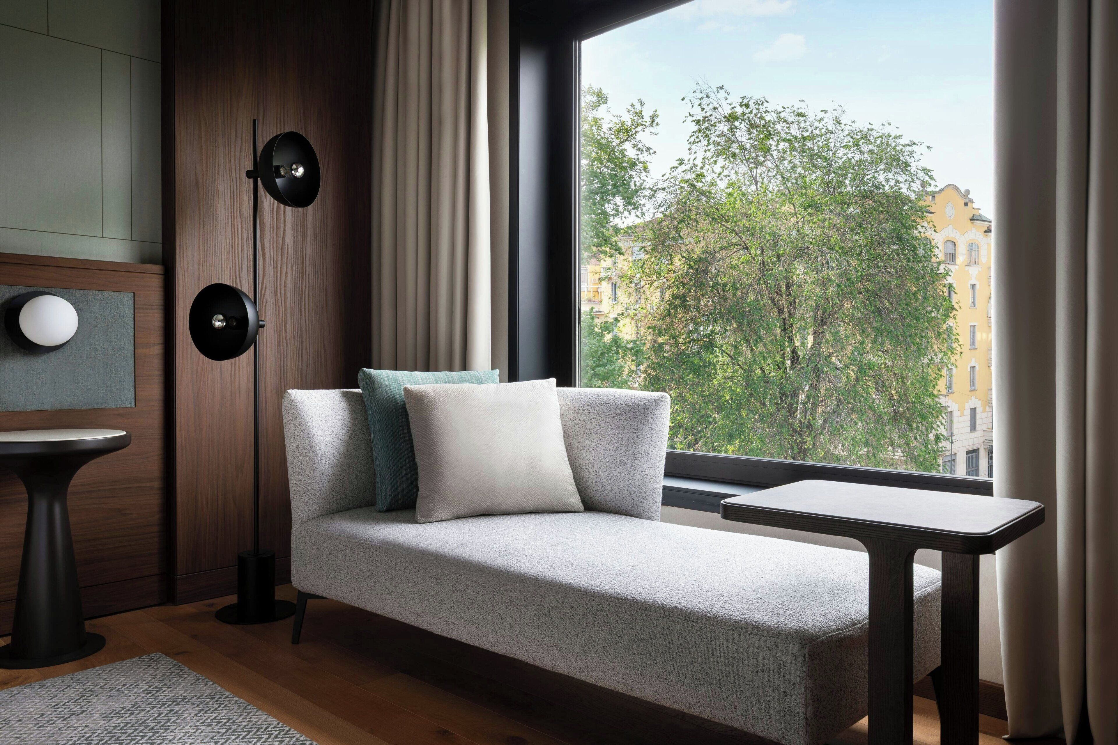 A modern living room with a sofa, a side table, a floor lamp, and a view of a tree-lined street through a large window.