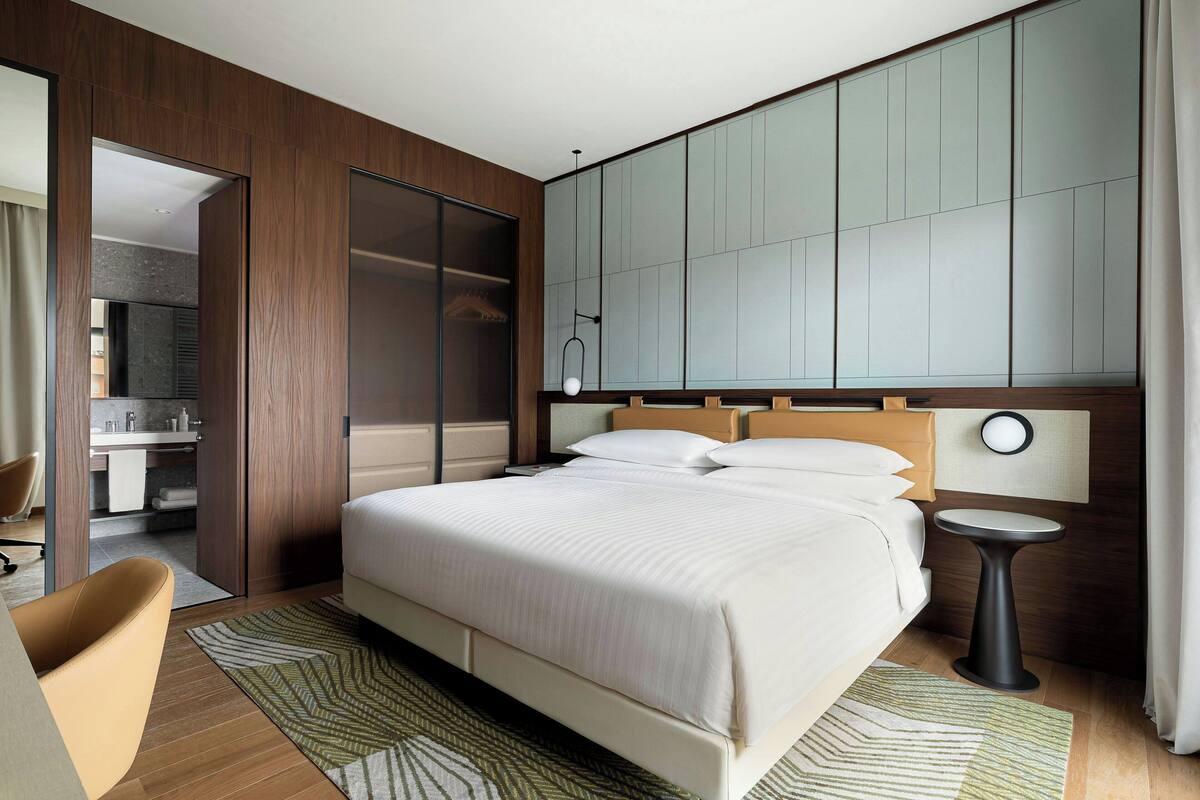 Quarto de hotel moderno com uma cama grande, uma mesa de cabeceira, uma cadeira e vista para o banheiro.