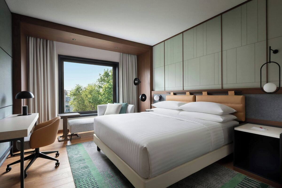 Quarto de hotel moderno com uma cama grande, uma escrivaninha, uma cadeira e vista para o exterior.