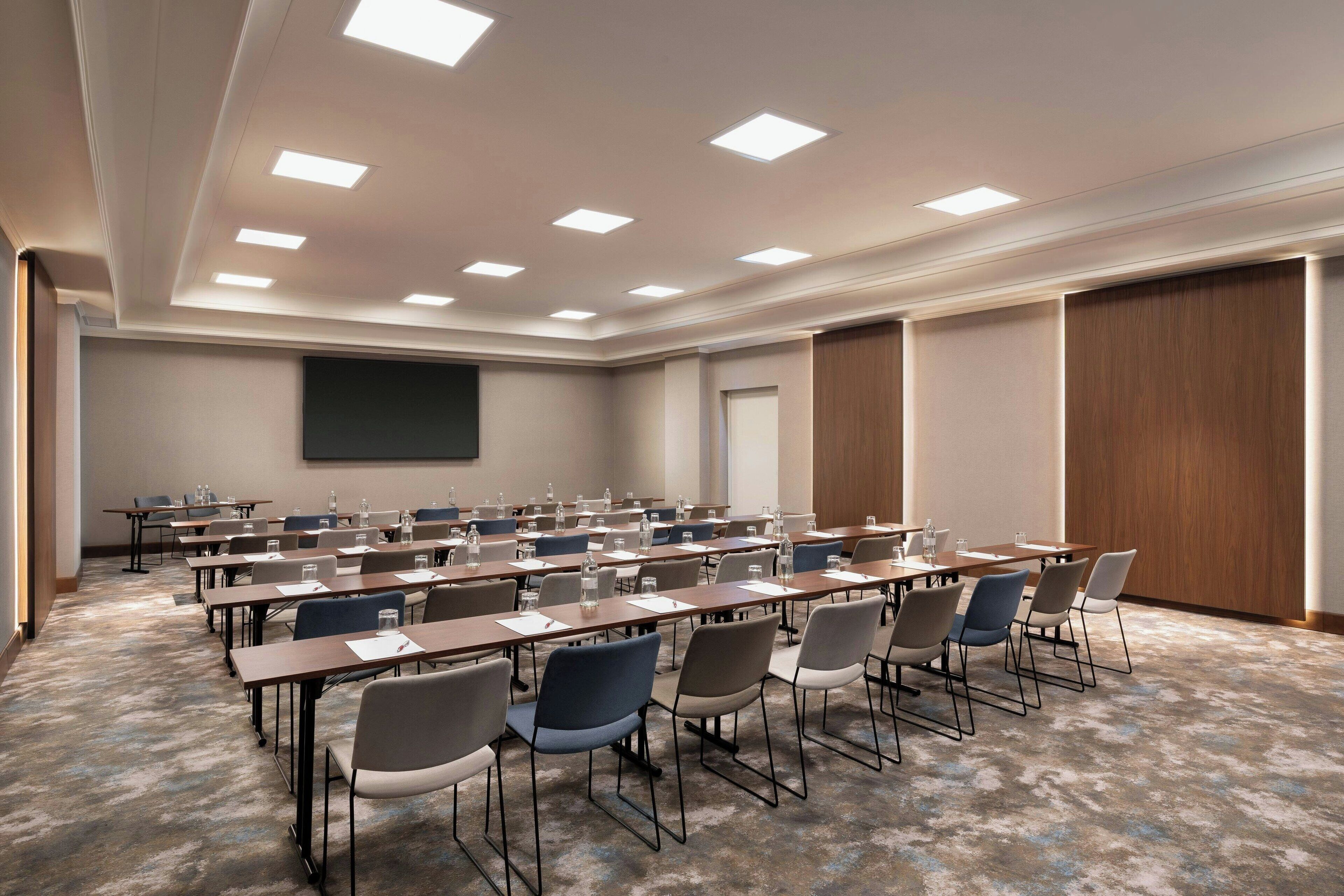 Un salón de conferencias con varias mesas y sillas, una pantalla grande y luces empotradas en el techo.