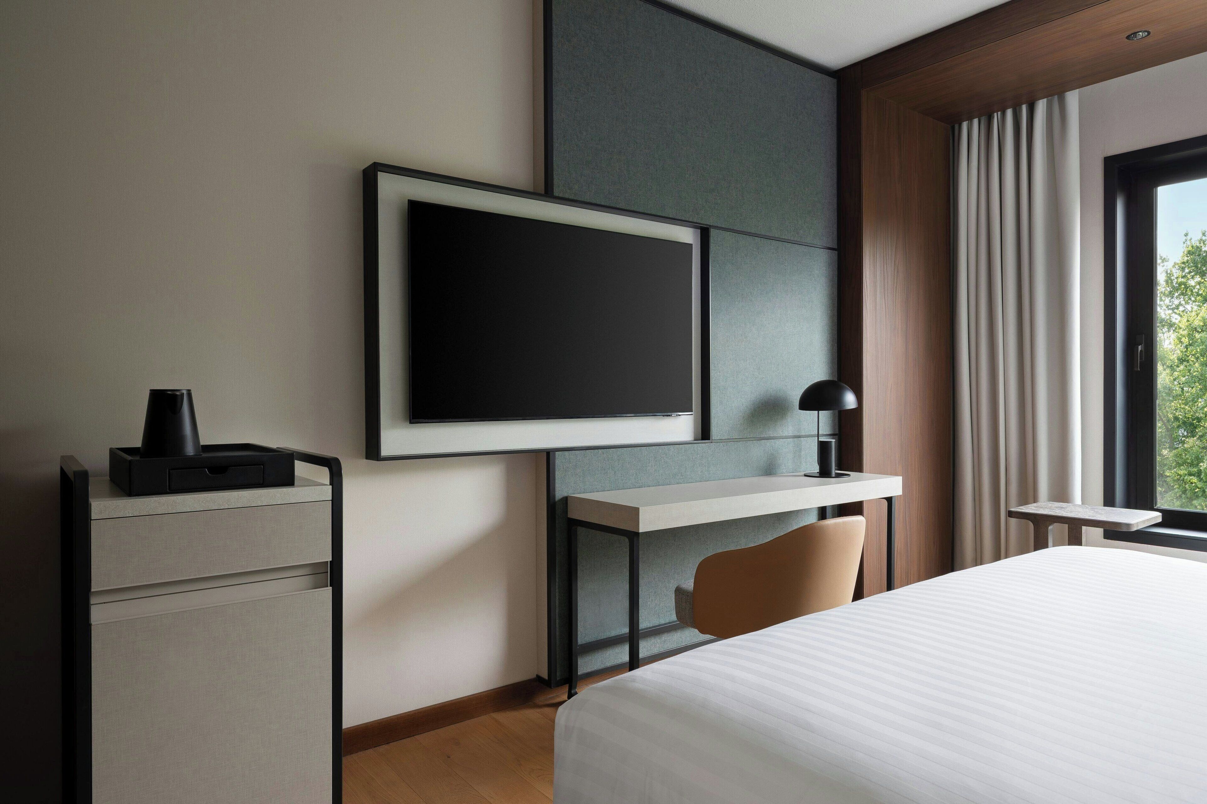 A hotel room with a bed, a desk with a chair, a TV mounted on the wall, and a window with curtains.