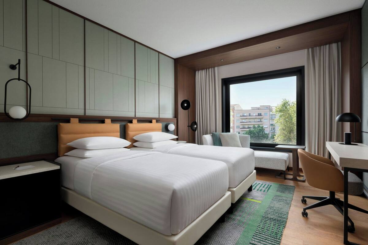 Quarto de hotel com duas camas, uma escrivaninha e uma grande janela com vista para a cidade.