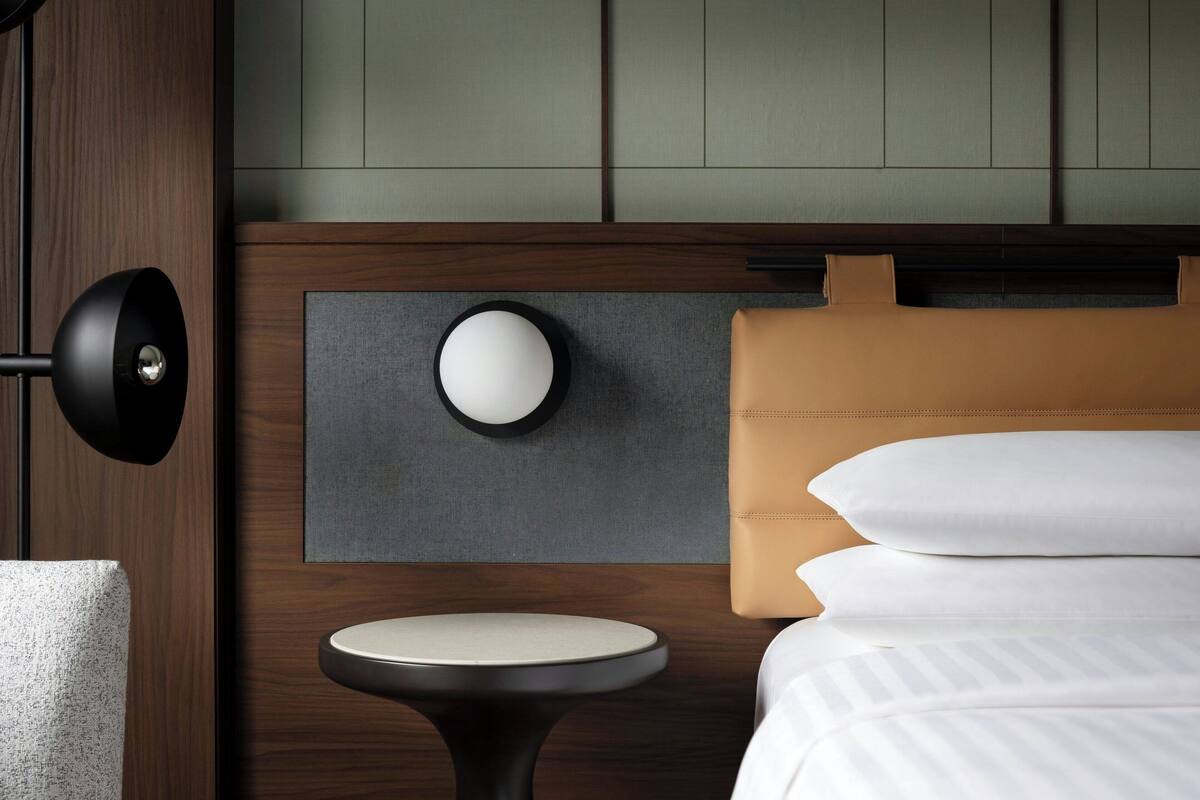 Quarto de hotel moderno com cabeceira de madeira, luminária redonda fixada na parede, uma pequena mesa redonda e cama com roupa de cama branca.