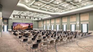 Un'ampia sala conferenze con file di sedie, un grande dipinto raffigurante un castello e un soffitto alto ornato da lampadari.