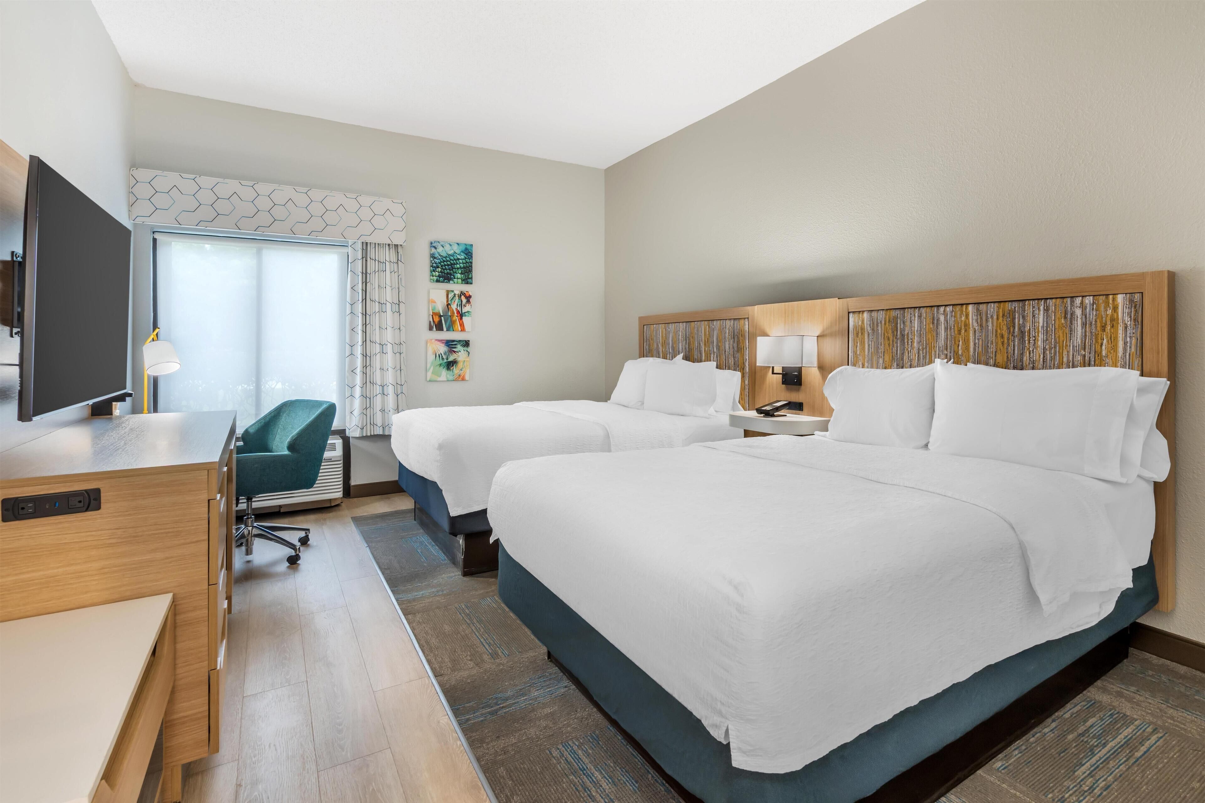 A hotel room with two beds, a desk, a chair, and a TV.