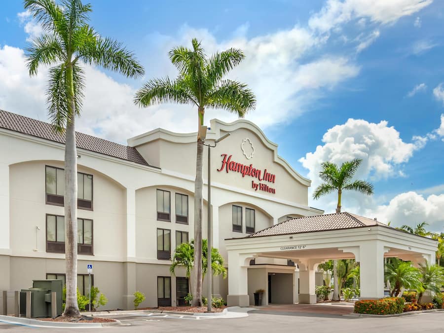A Hampton Inn by Hilton hotel with a clear sky, palm trees, and a covered entrance.