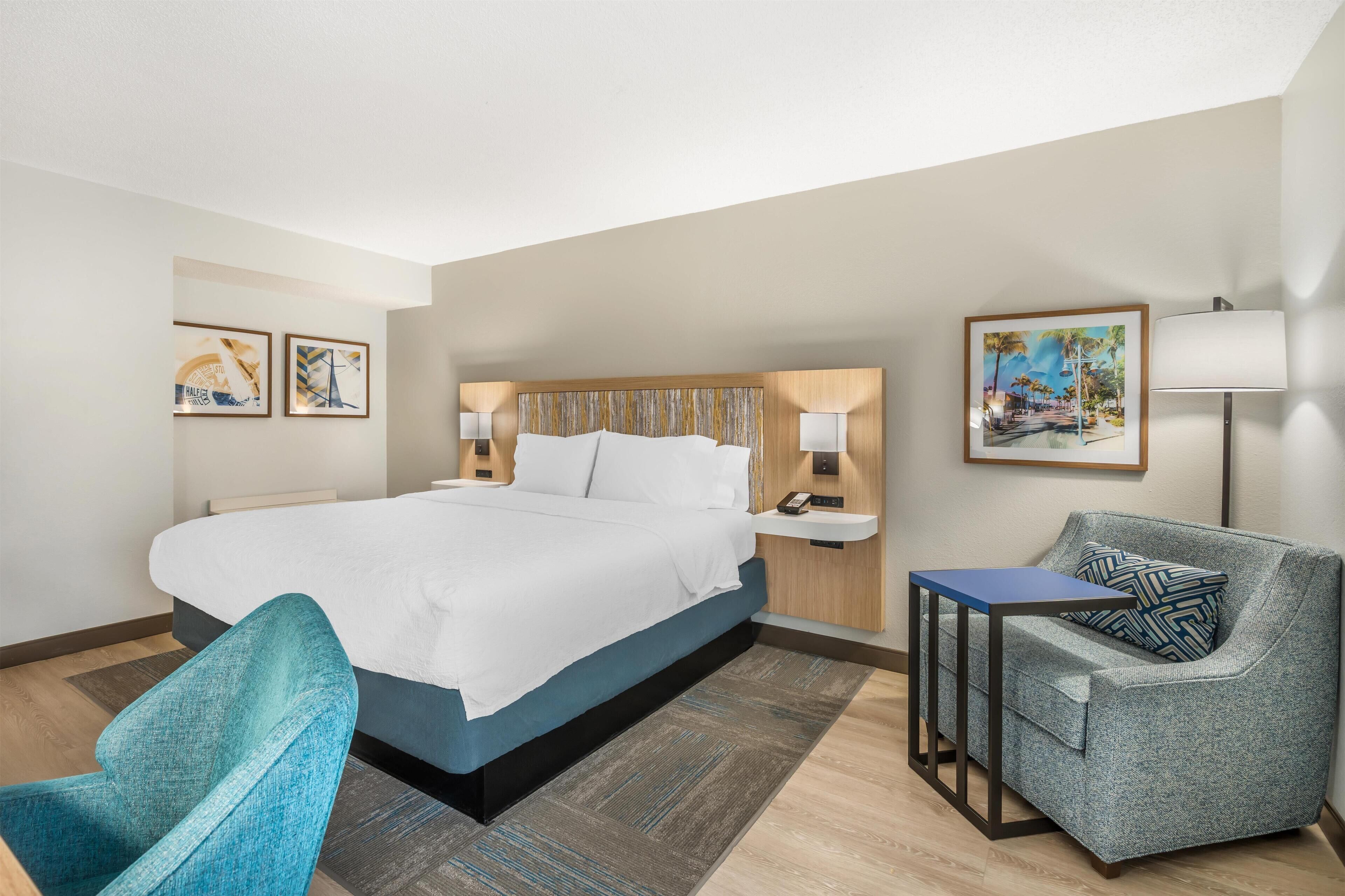 A hotel room with a large bed, a seating area with a chair and a table, and artwork on the walls.