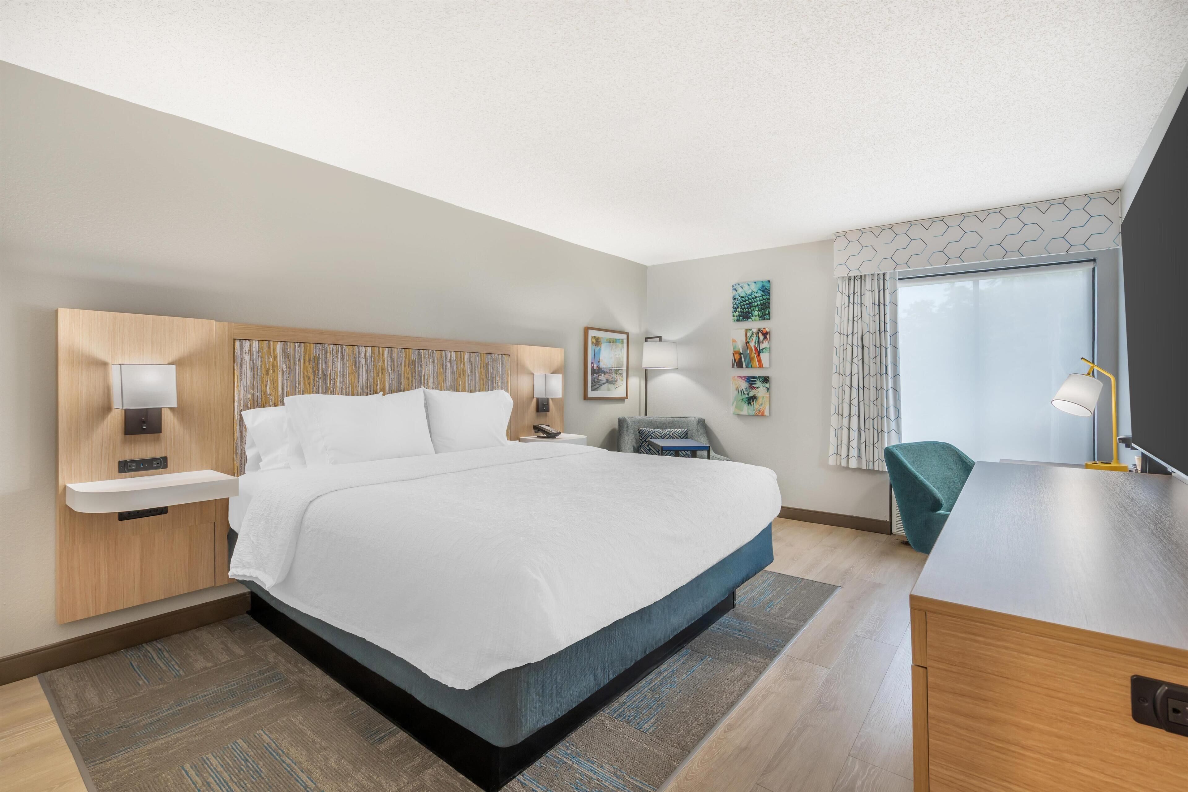 Une chambre d’hôtel moderne avec un grand lit, des tables de chevet, un bureau et une fenêtre avec des rideaux.