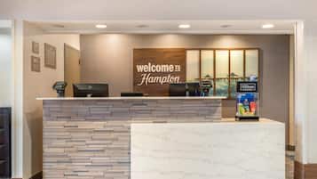 A hotel reception desk with a 'Welcome to Hampton' sign, a digital check-in station, and a vending machine.