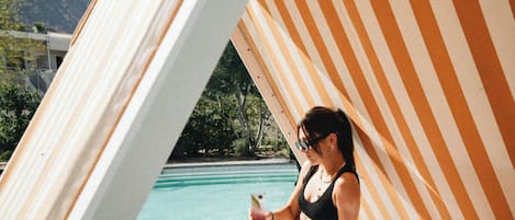 A person sitting on a lounge chair by a pool with a striped awning, a clear view of the pool, and mountains in the background.