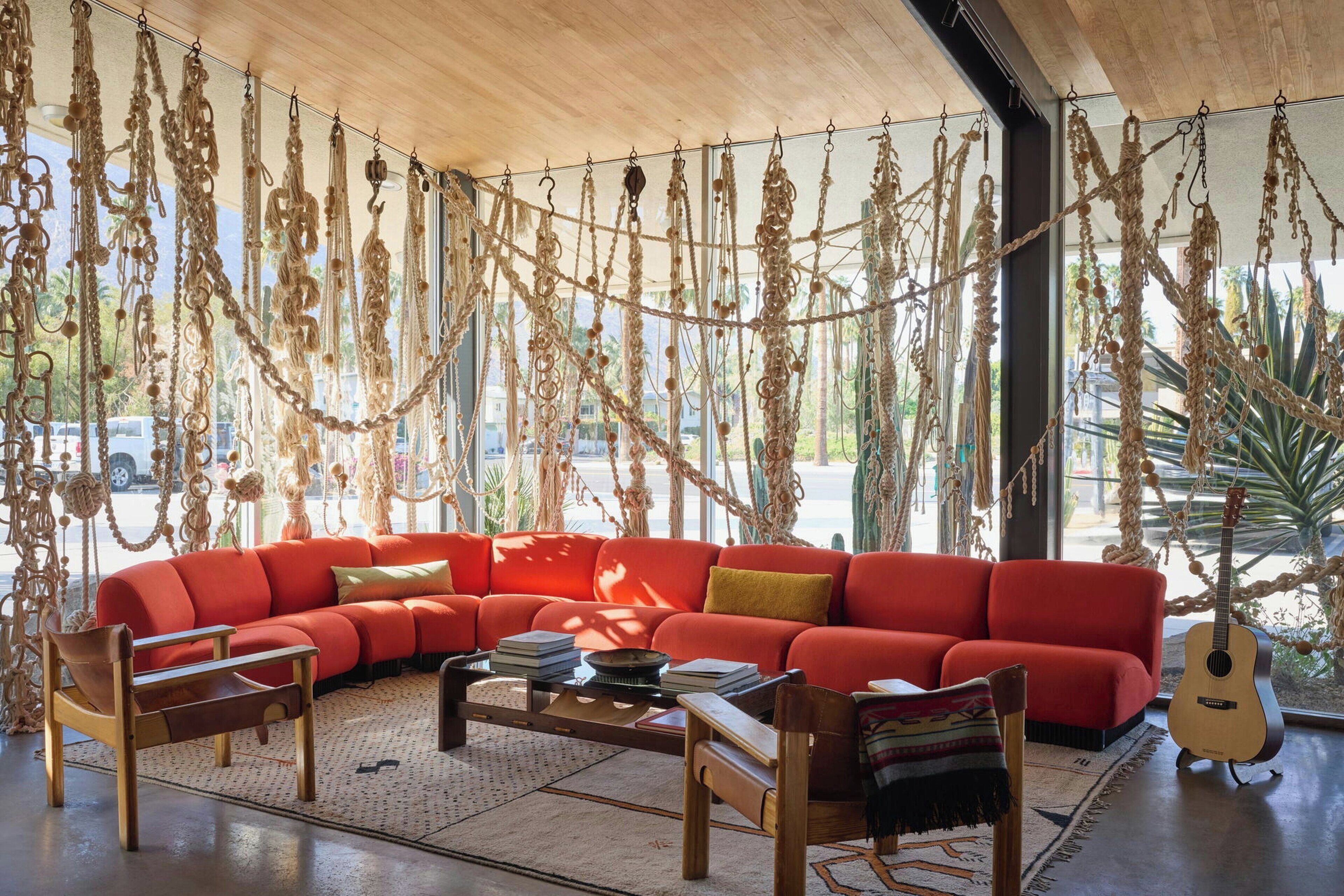 Una sala moderna con un sofá rojo, una mesa de centro de madera y sillas, decorada con tapices de macramé y una guitarra.