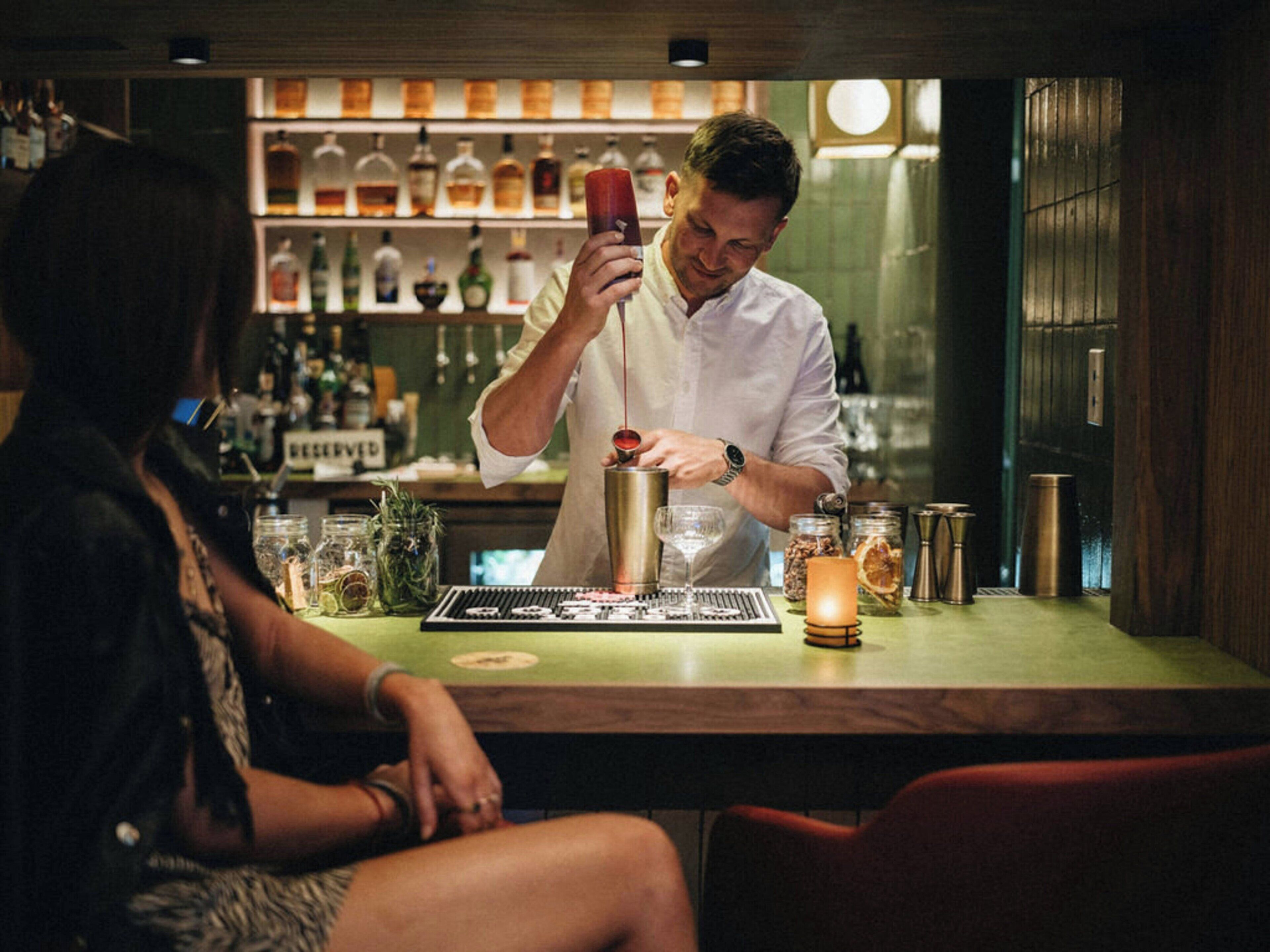 A bar setting with a bartender preparing a drink, a bar counter with various bottles and a menu, and a seated customer.