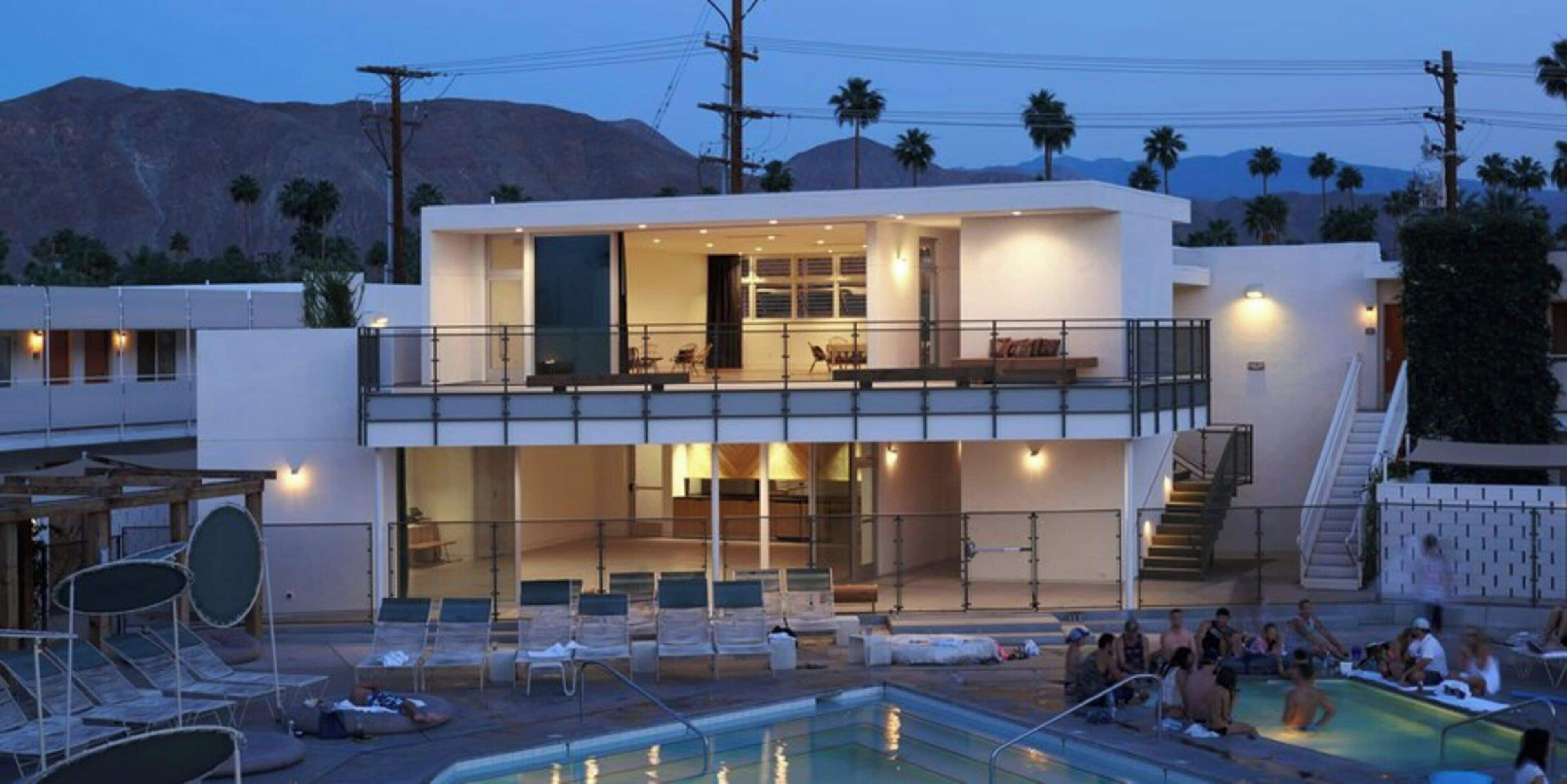 A modern two-story building with a pool area, outdoor seating, and a view of mountains.