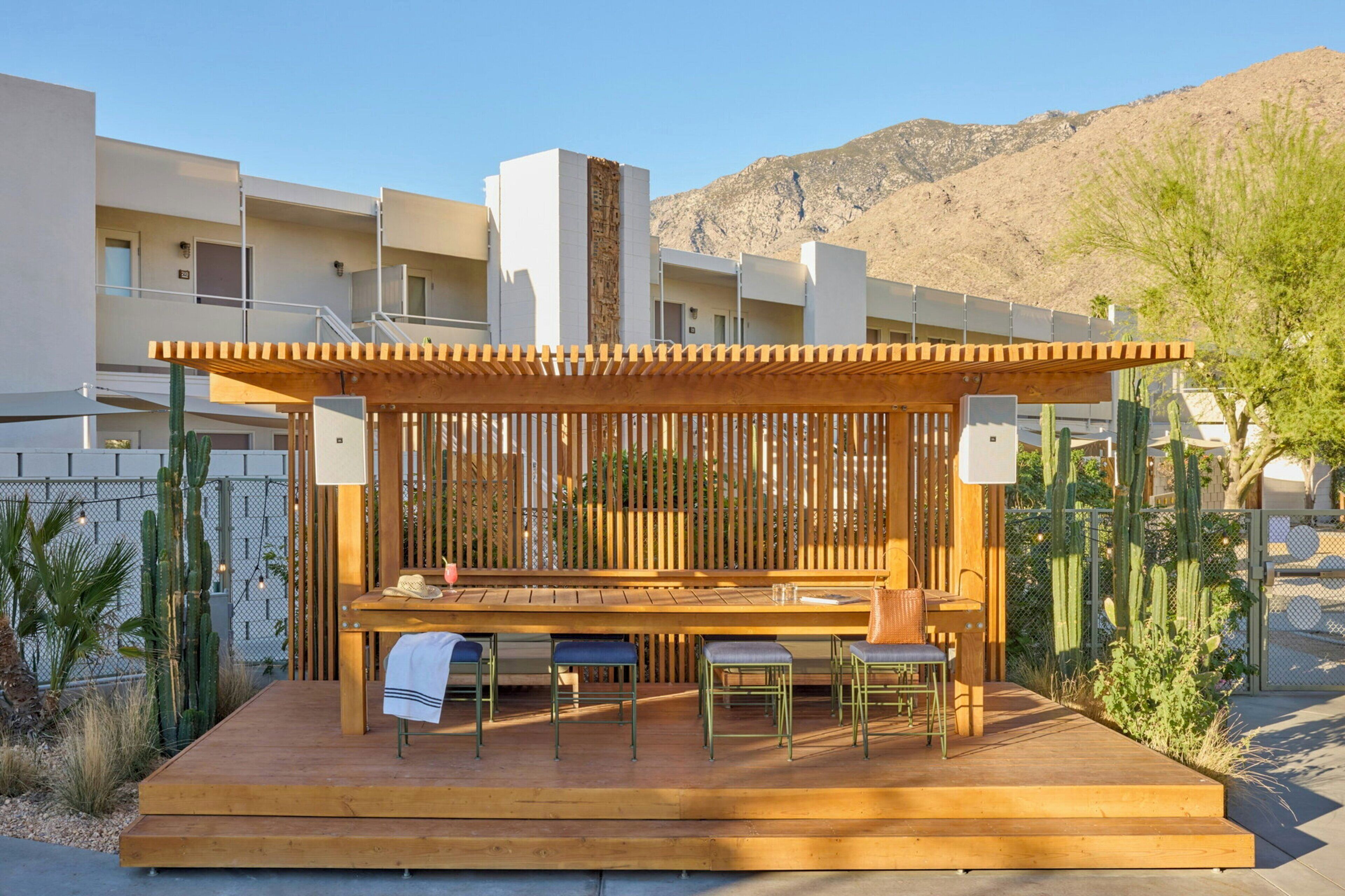 An outdoor wooden deck with a table and chairs, surrounded by a wooden fence, in a residential area with buildings and cacti.