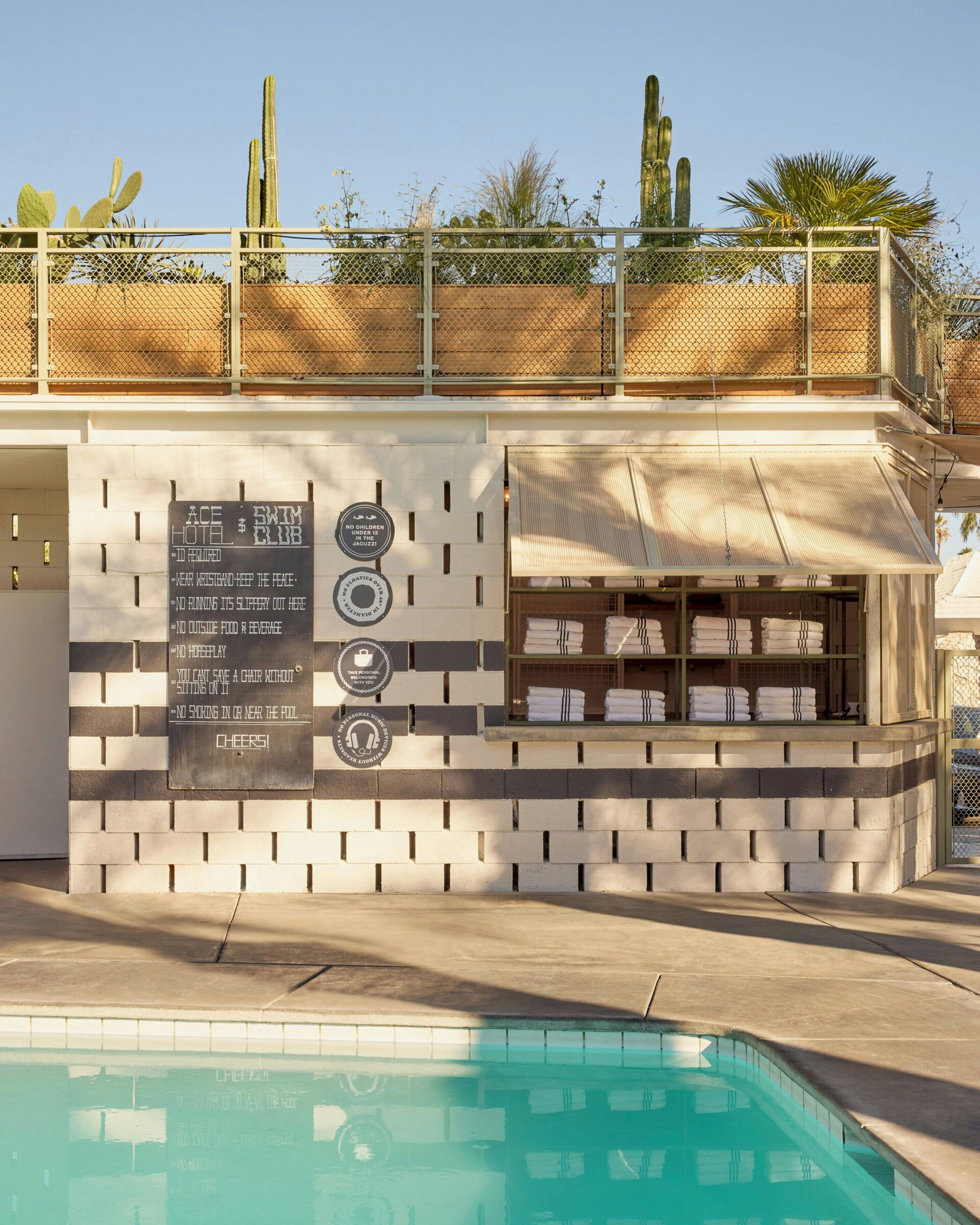 A poolside area with a sign listing hotel rules, a bar with towels, and cacti in the background.