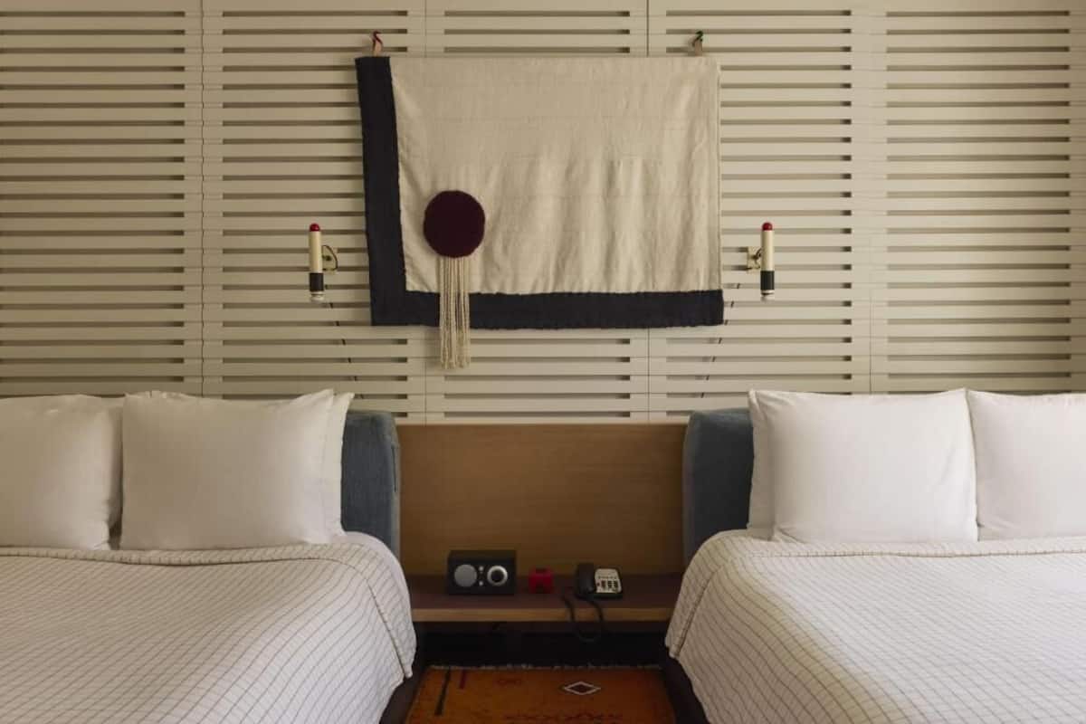 A hotel room with two beds, a wooden headboard, a small table with a radio and phone, and a wall-mounted decorative item.
