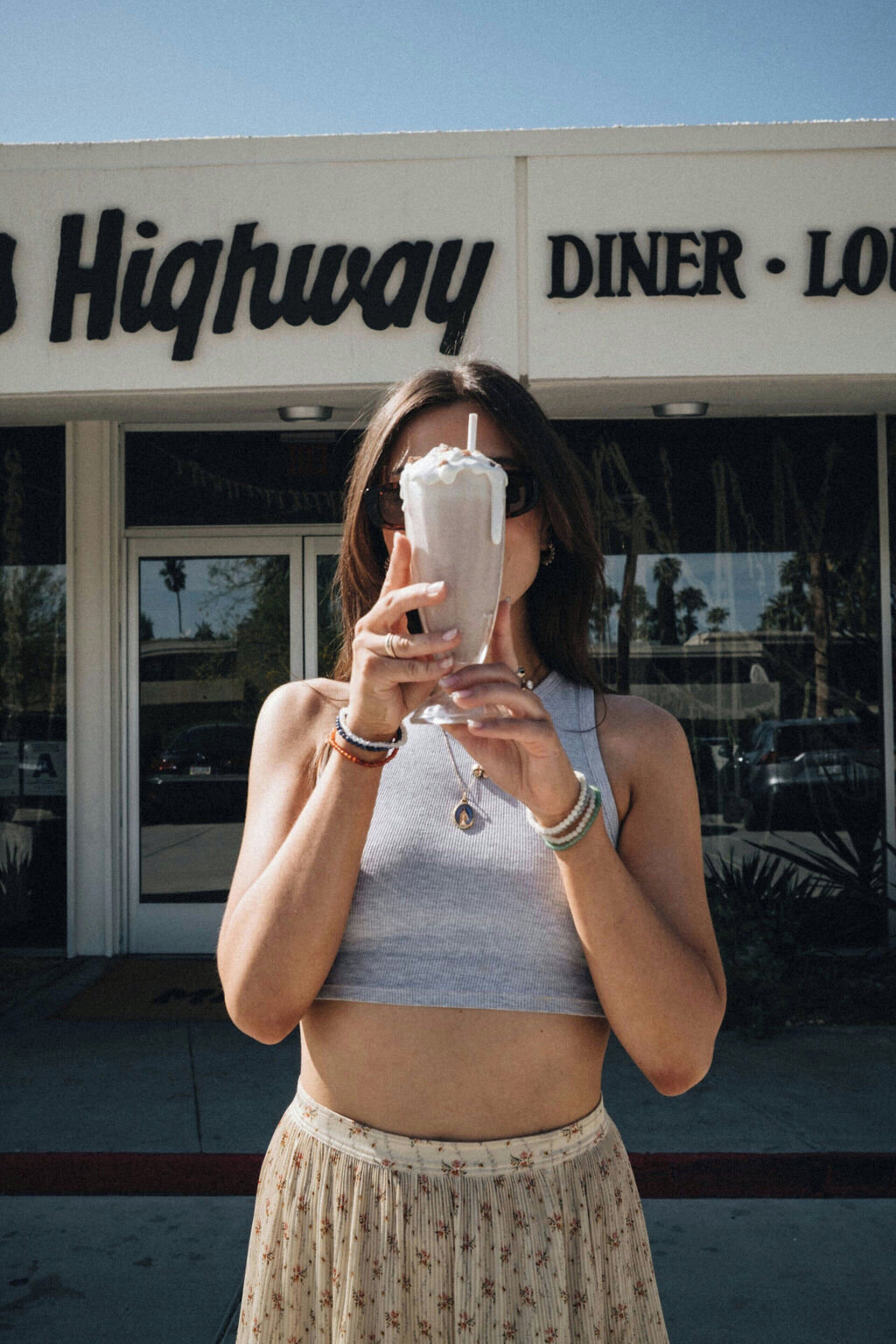 A person holding a drink in front of a building with a sign that reads 'Highway Diner - Lou.'