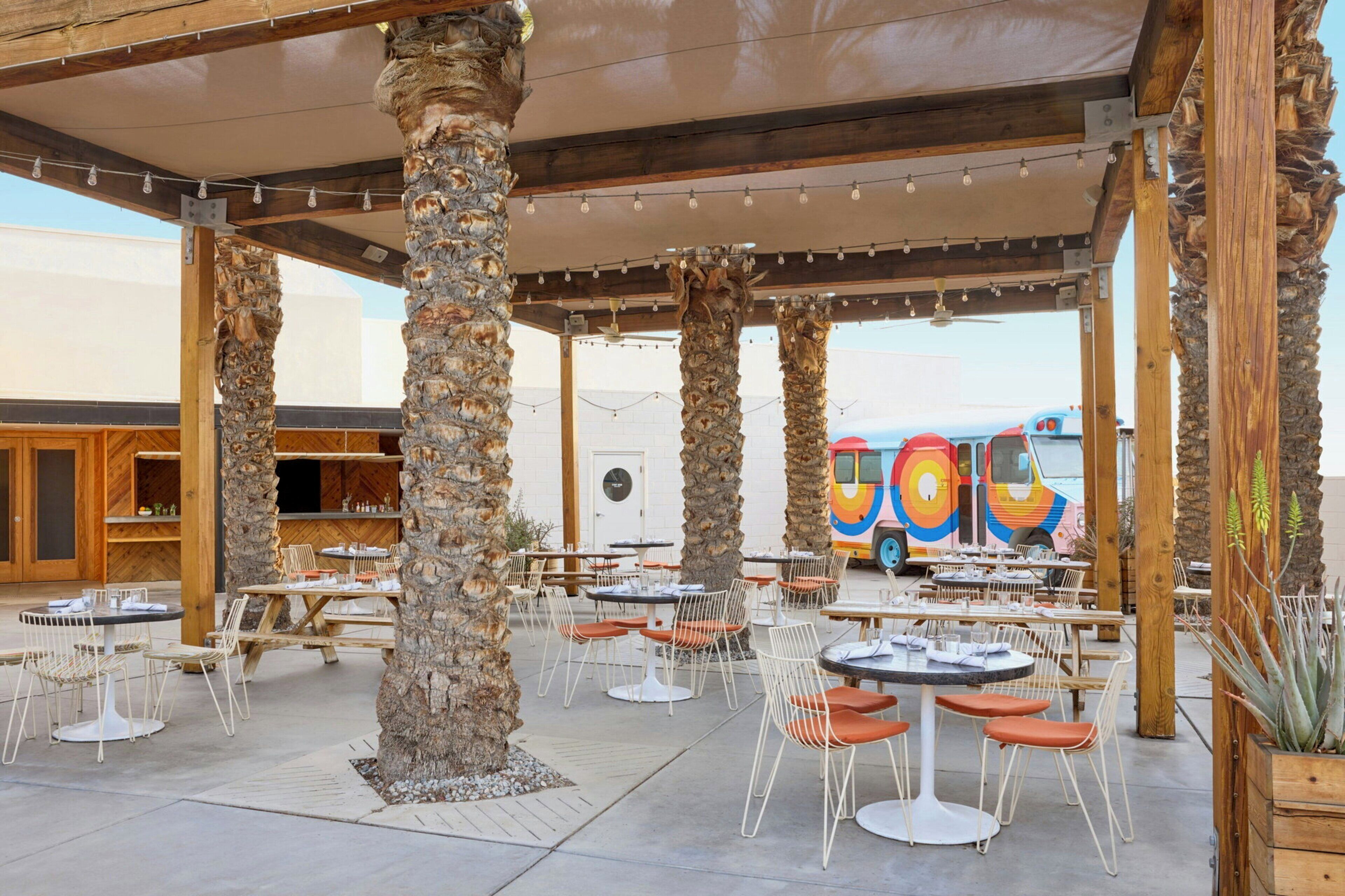 Un espace de restauration en plein air avec des tables et des chaises, des palmiers et un autobus coloré en arrière-plan.