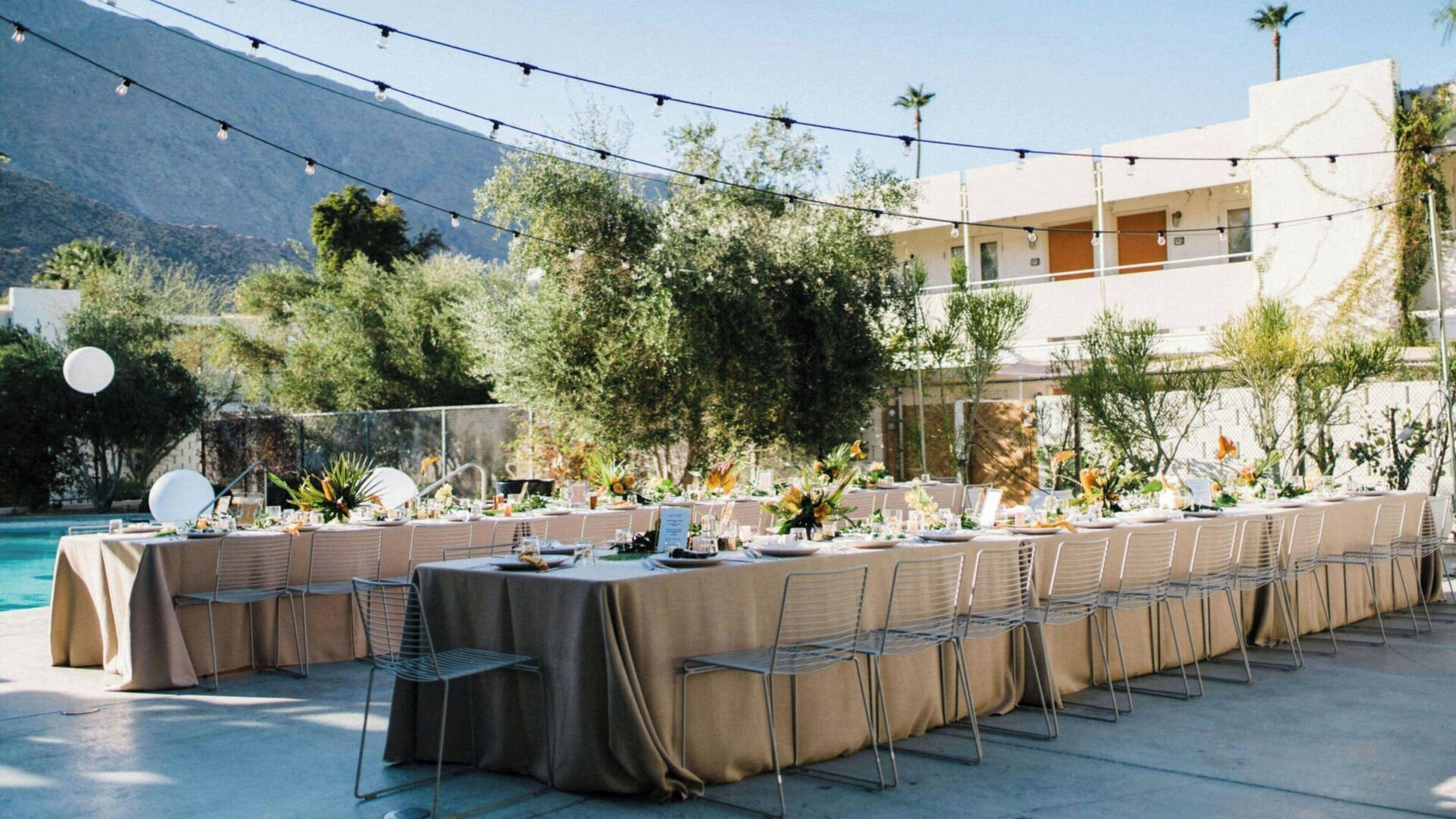 An outdoor dining area with tables set for a meal, surrounded by greenery and a pool.