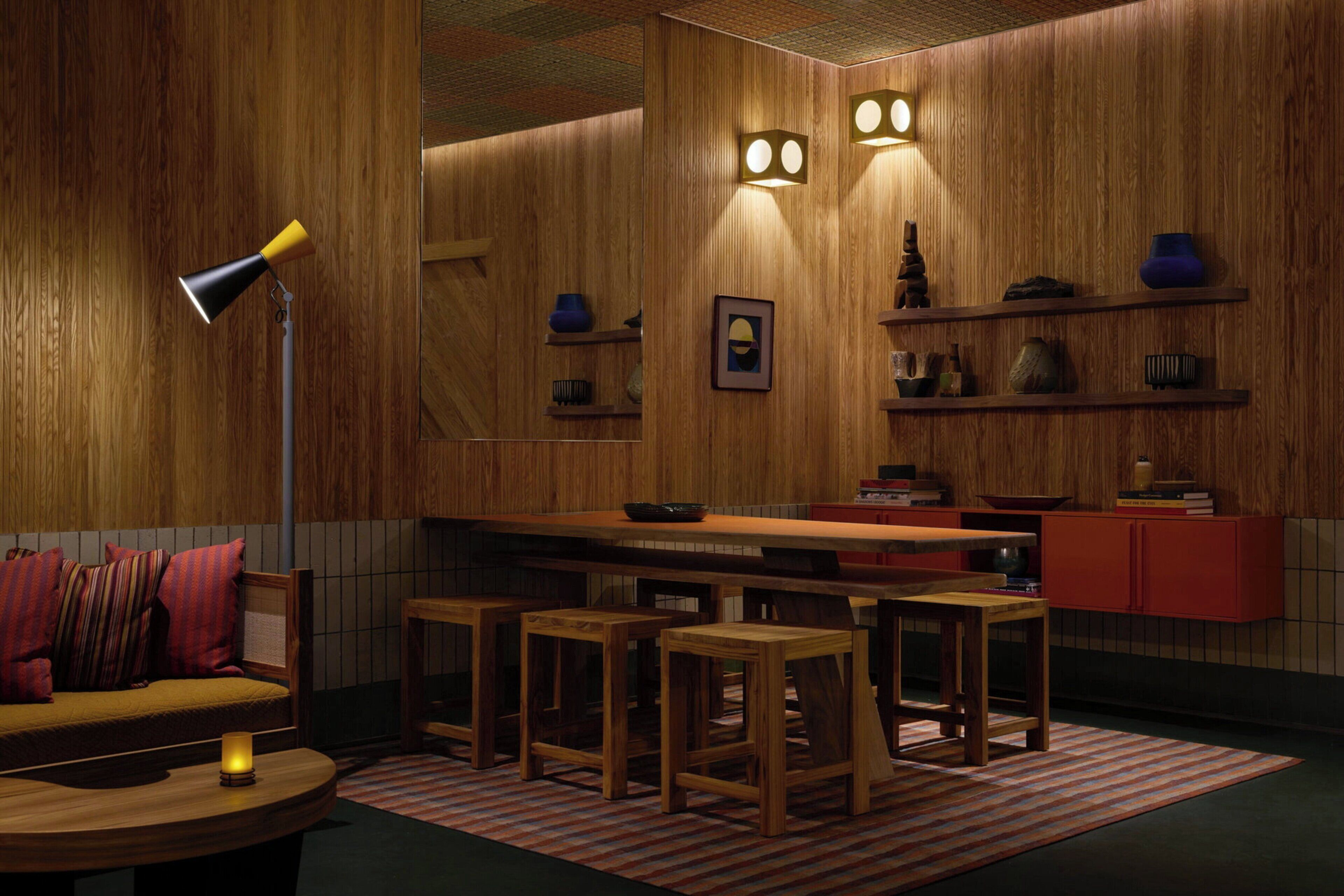 A cozy room with a sofa, a table, a lamp, a kitchen area with stools, and shelves with various items.