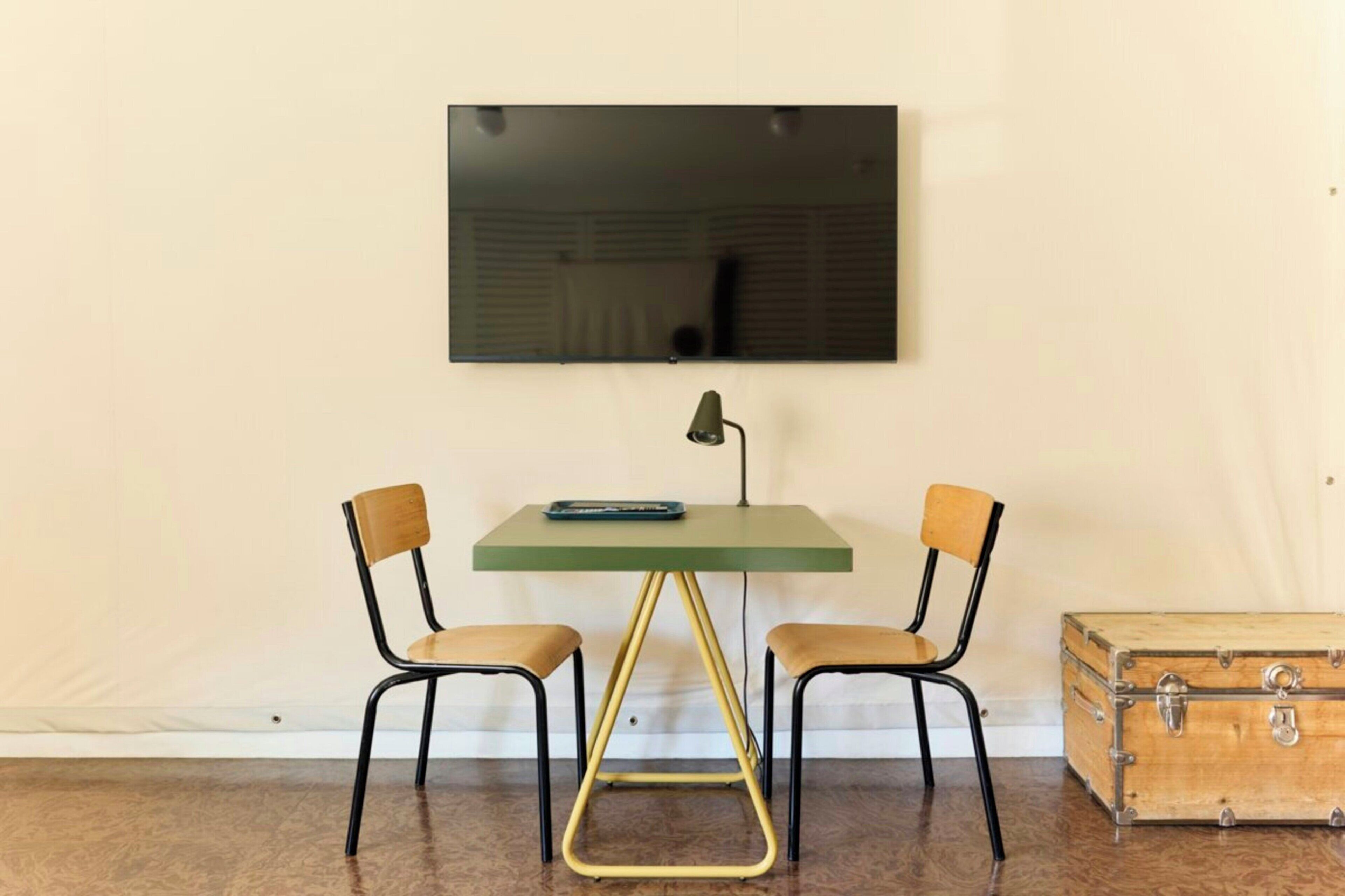 Um quarto pequeno com uma mesa verde, duas cadeiras, uma televisão na parede e uma mala antiga.