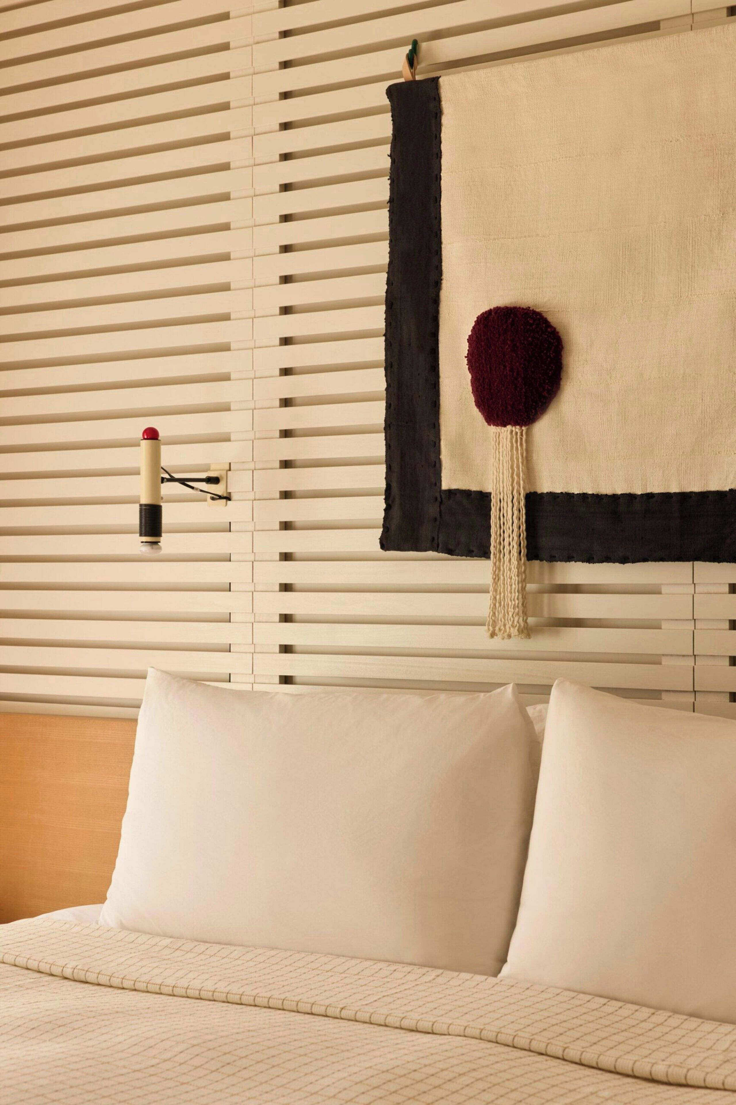 Un lit avec une literie blanche, un mur avec des stores horizontaux et un objet décoratif fixé au mur.