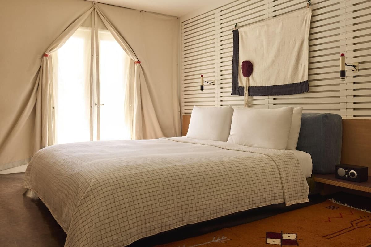 A bedroom with a bed, curtains, a wall-mounted radio, and a window with blinds.