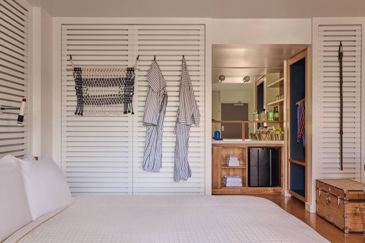 A bedroom with a bed, a wardrobe with hanging clothes, a mirror, and a small wooden cabinet with bottles.