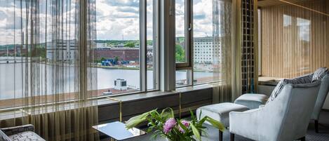 A modern hotel room with large windows, a sofa, a chair, and a small table with a vase of flowers.