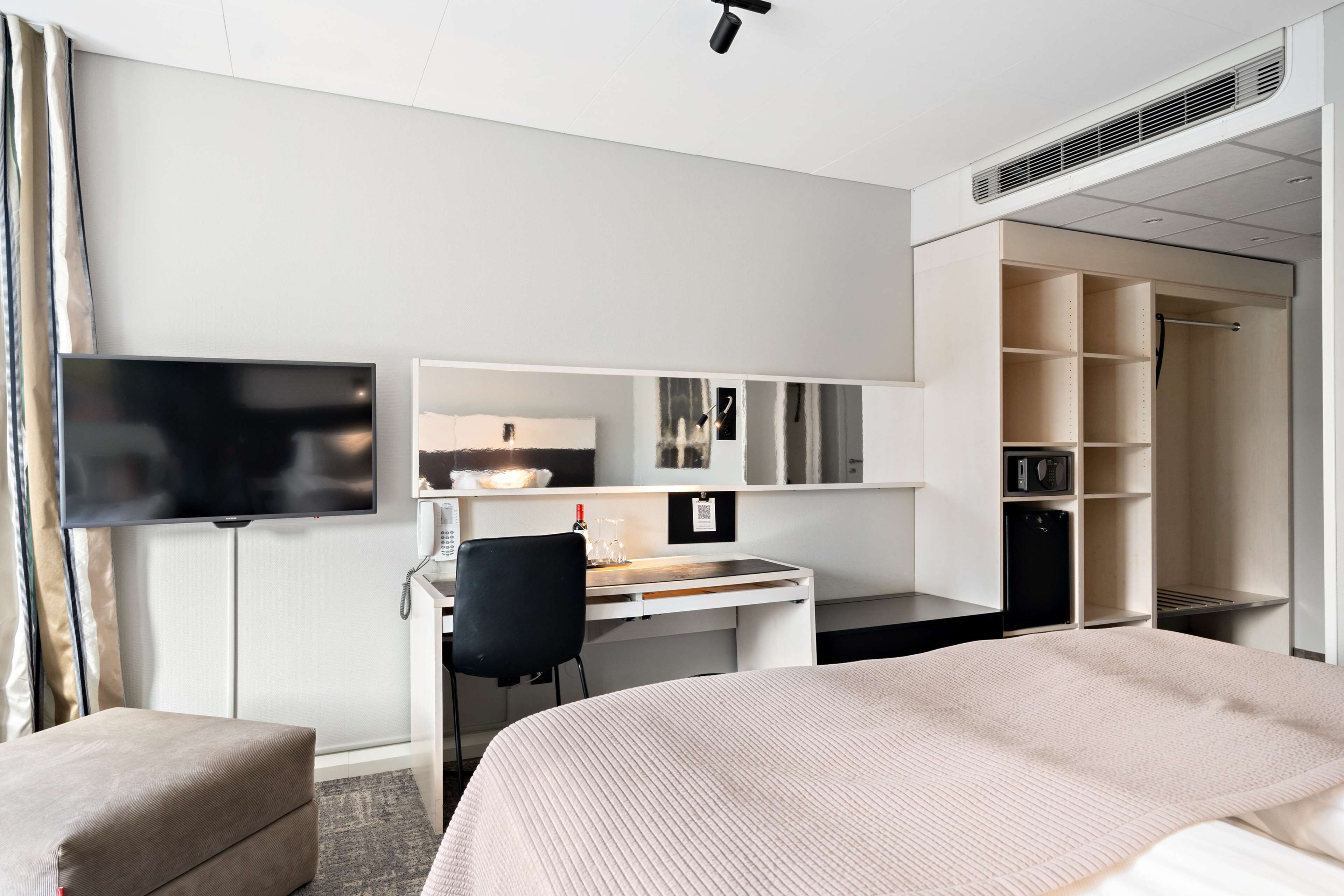 Una habitación de hotel con una cama, un escritorio con silla, un televisor, un armario y un espejo.