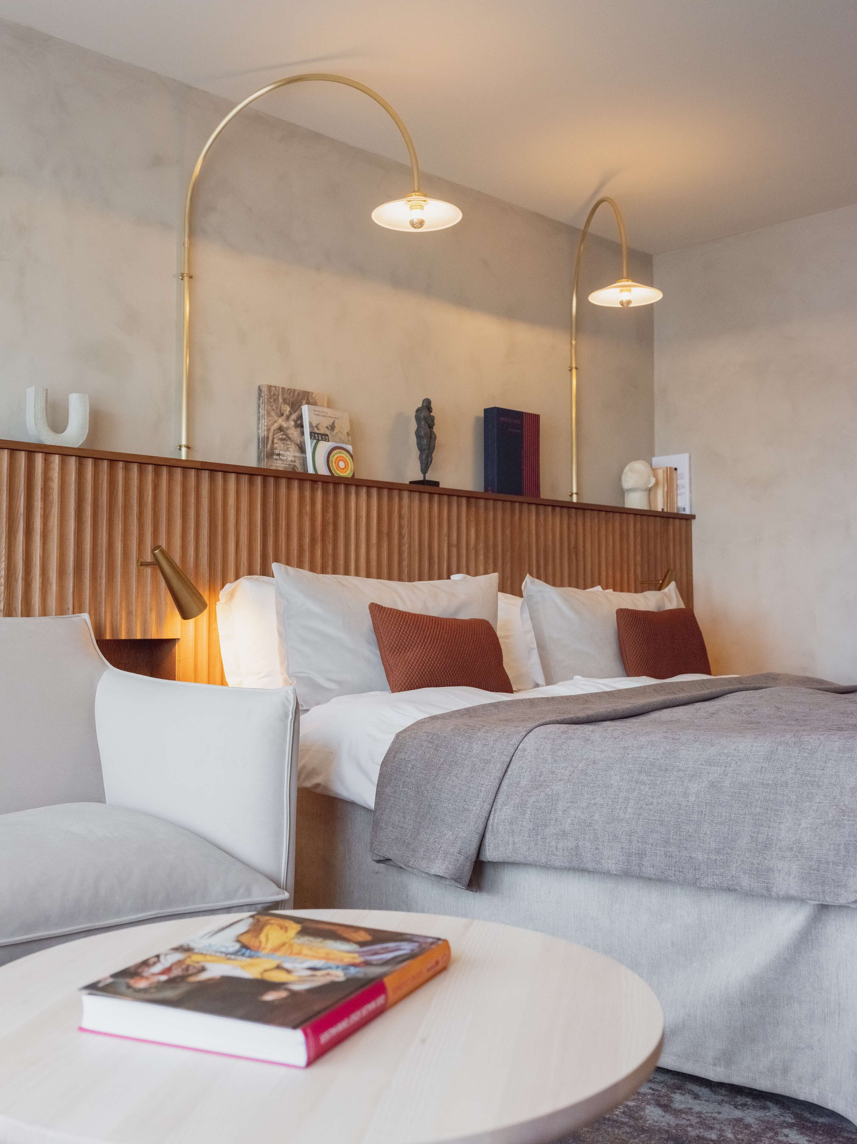 A modern hotel room with a bed, a sofa, a round coffee table with a book, and two wall-mounted lamps.