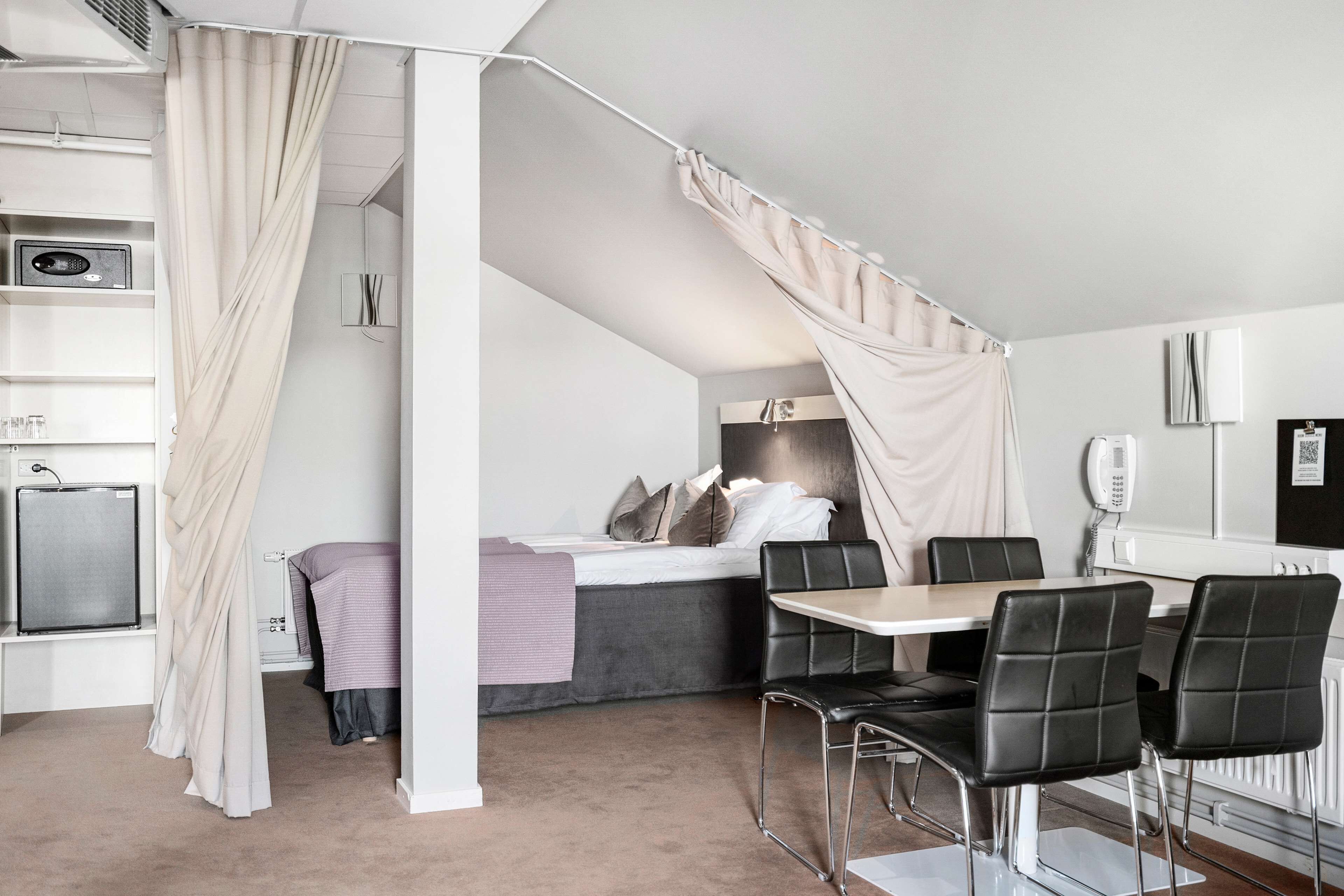Quarto moderno com uma cama, uma escrivaninha e uma cadeira. Há uma janela com cortinas, um radiador e uma estante com objetos.