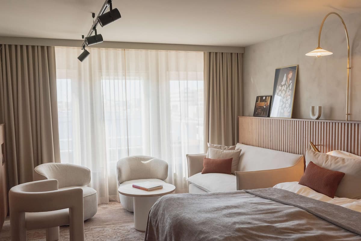 Una habitación de hotel moderna con una cama, dos sillones, una pequeña mesa redonda y un sofá. Cuenta con un ventanal con cortinas translúcidas, un cuadro colgado en la pared y una lámpara en el techo.