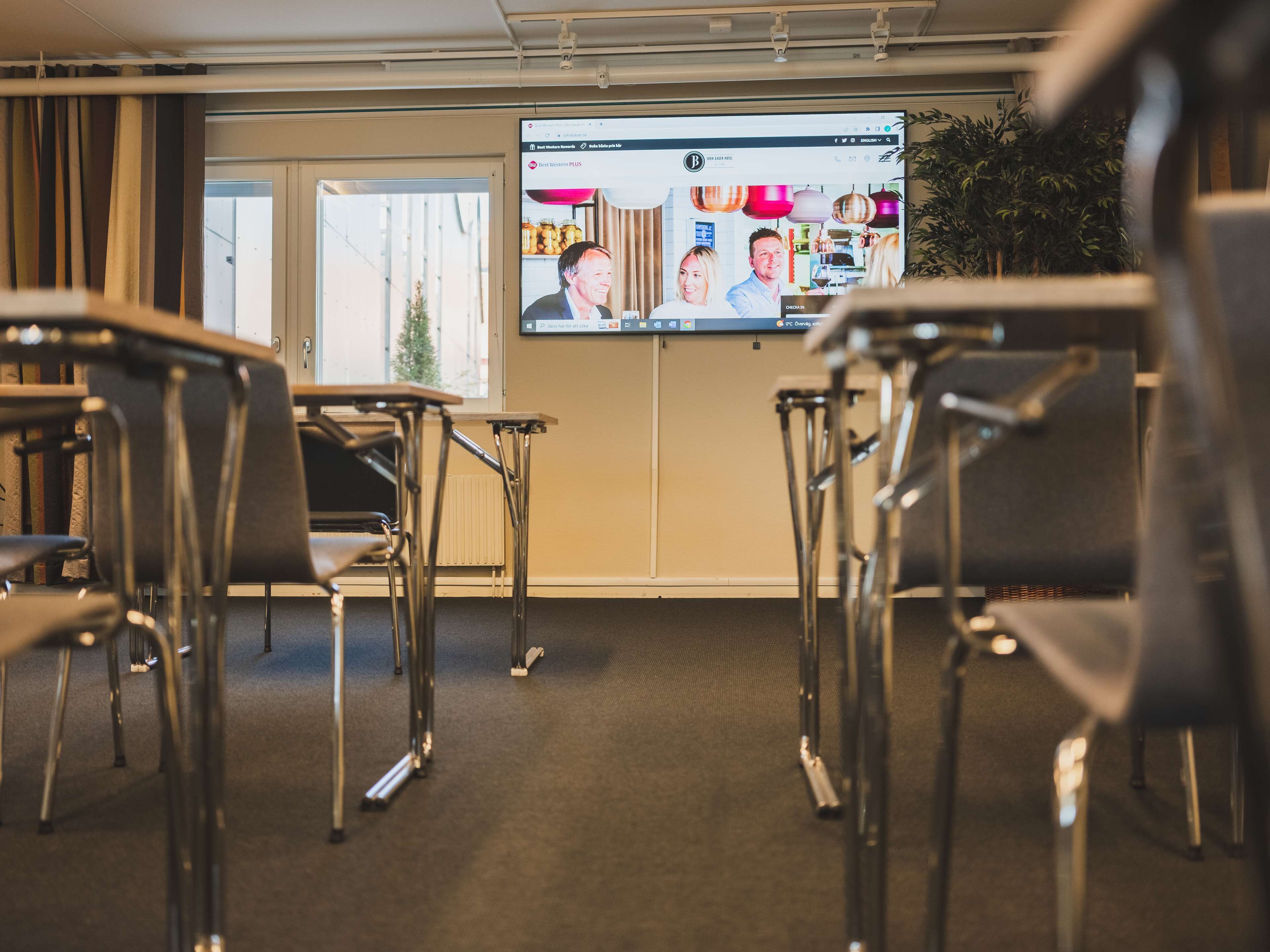 A conference room with metal-framed chairs, a large screen displaying a video, and a window with curtains.