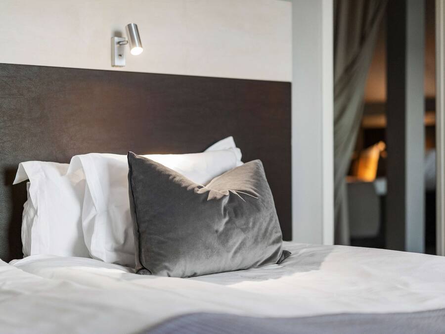 Una cama bien hecha con almohadas blancas y grises, un cabecero moderno y una lámpara de pared.