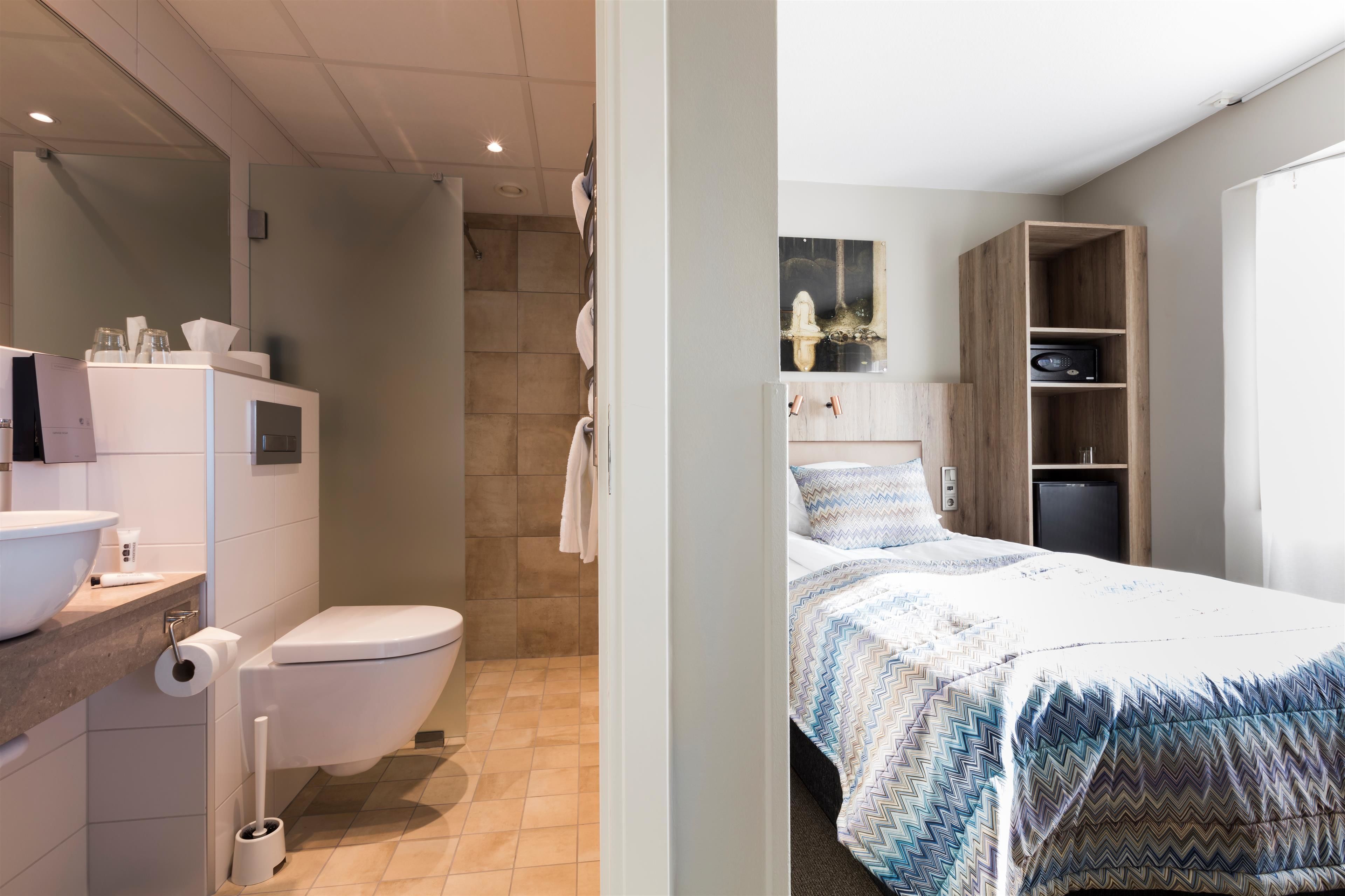Un baño moderno con inodoro, lavabo y ducha. La habitación cuenta con una cama con cubrecama estampado y un armario de madera.