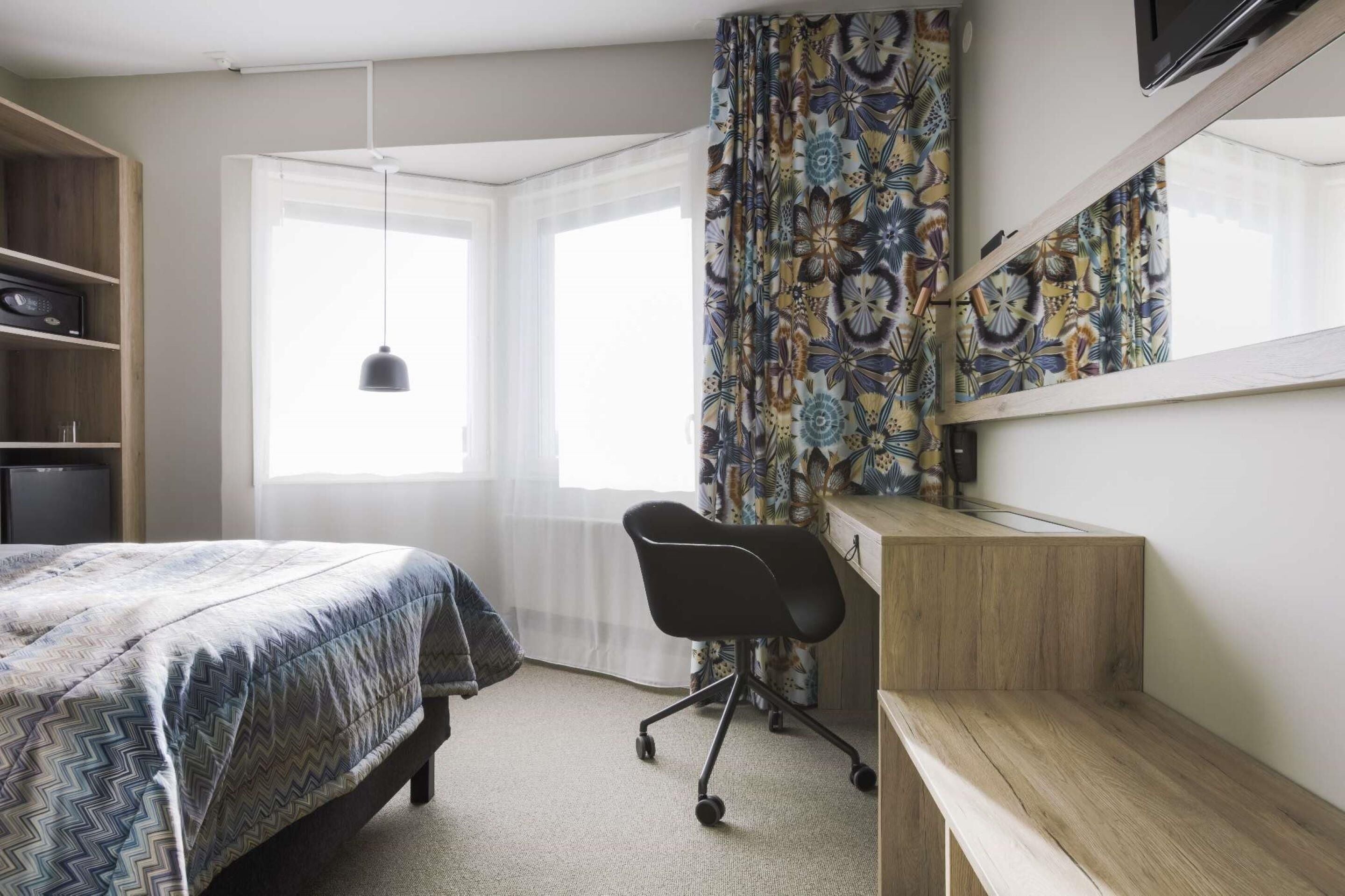A bedroom with a bed, a desk, a chair, a wardrobe, and a window with curtains.