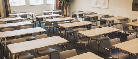 A classroom with multiple desks and chairs, large windows, and a plant.