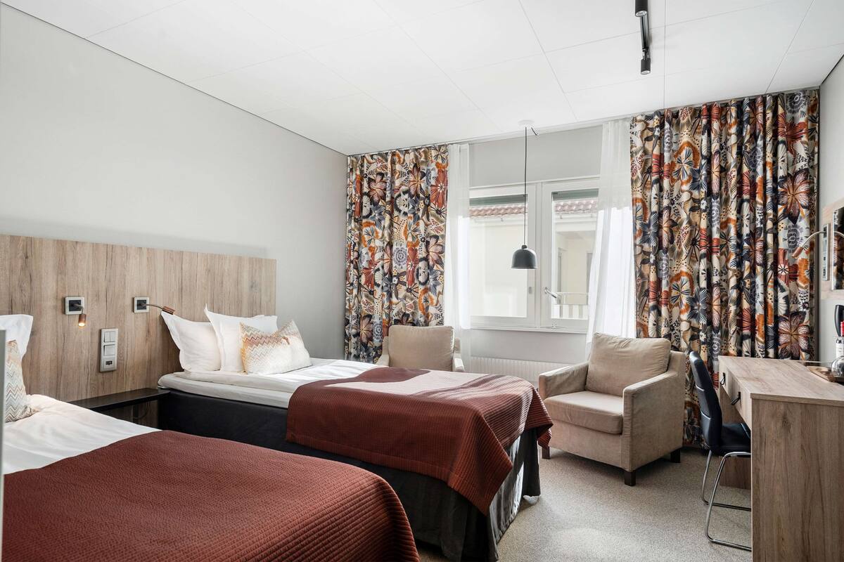 Habitación de hotel con dos camas, un escritorio, una silla y un ventanal con cortinas.