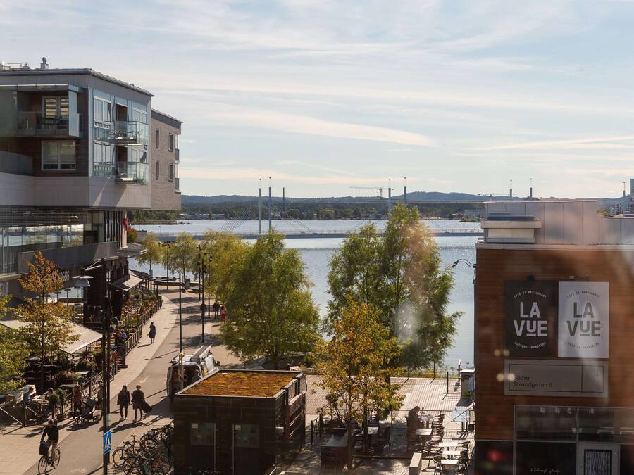 En utsikt över floden med moderna byggnader, ett kafé som heter ”La Vue” och en gångväg för fotgängare.