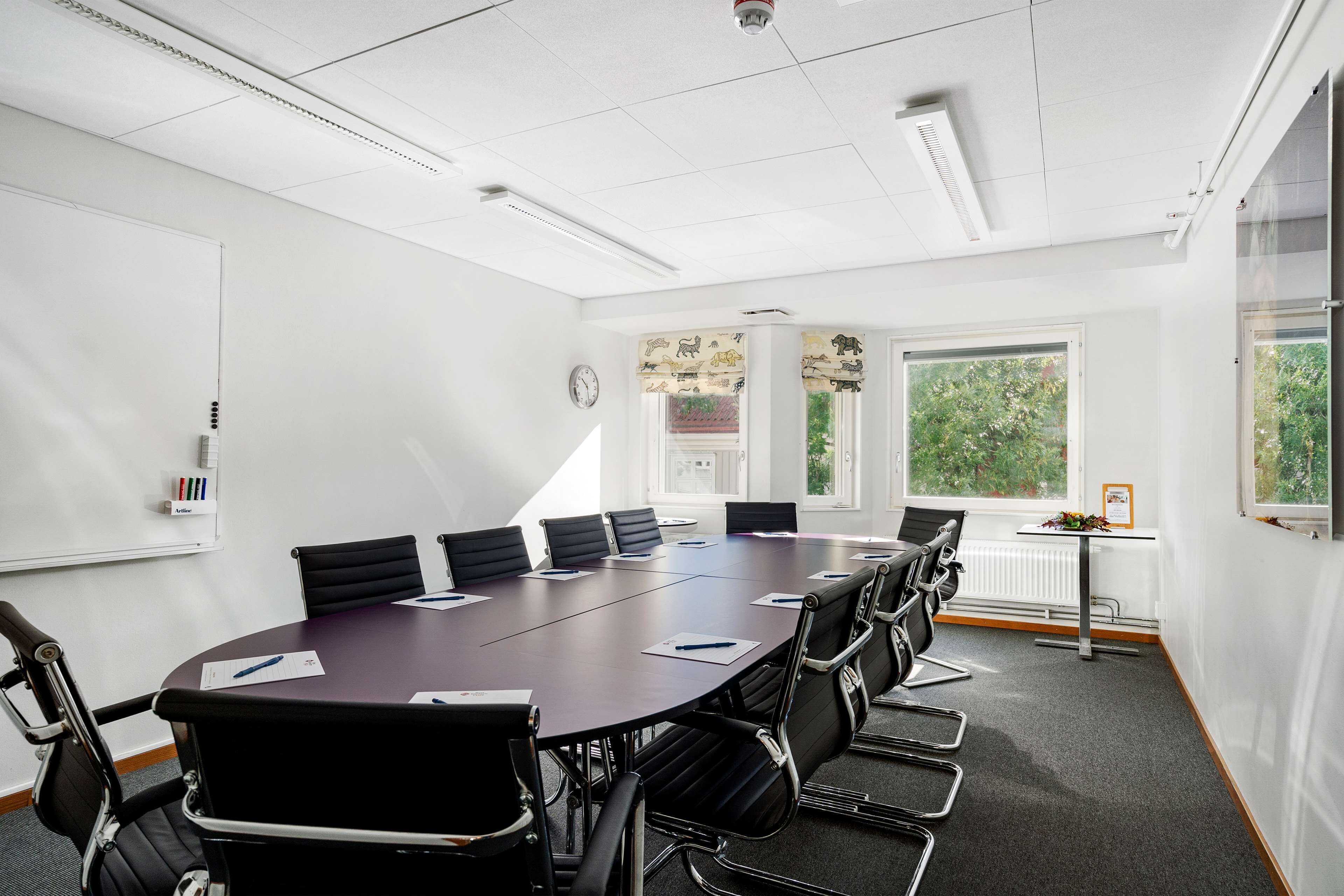 A conference room with a large table, black chairs, white walls, and windows with curtains.