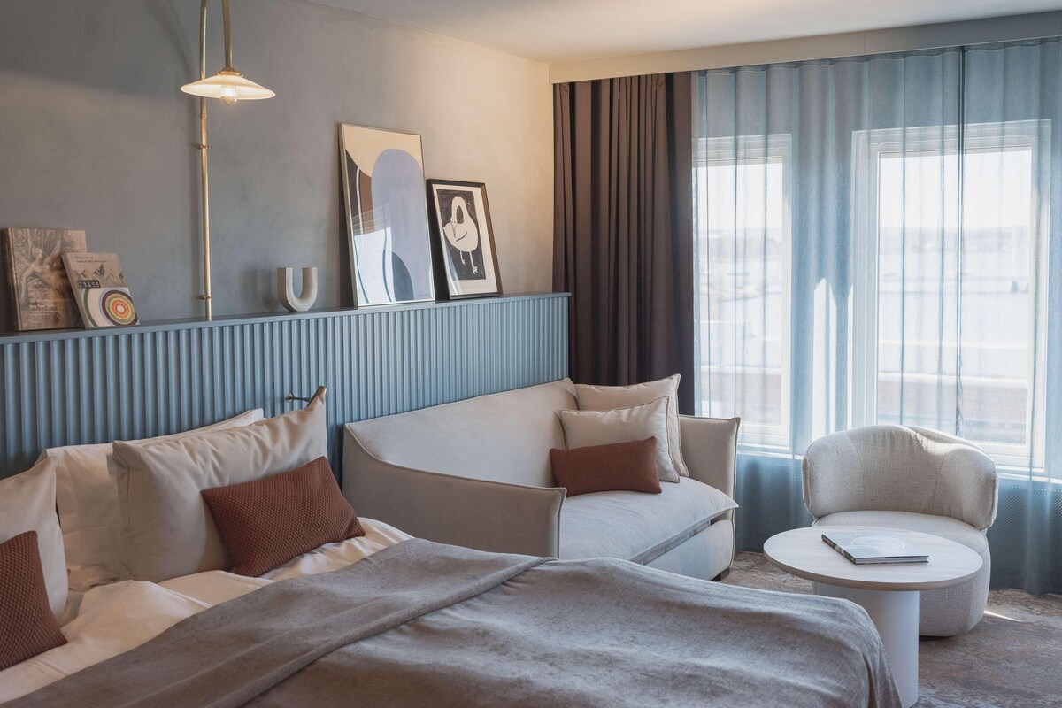 A modern hotel room with a bed, a sofa, a small round table, and a large window with curtains.