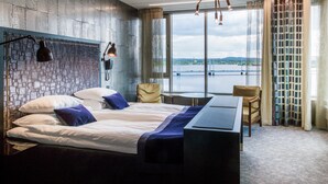 A modern hotel room with a large bed, a desk, and a view of a body of water through large windows. - Best Western Plus John Bauer Hotel (Jonkoping)