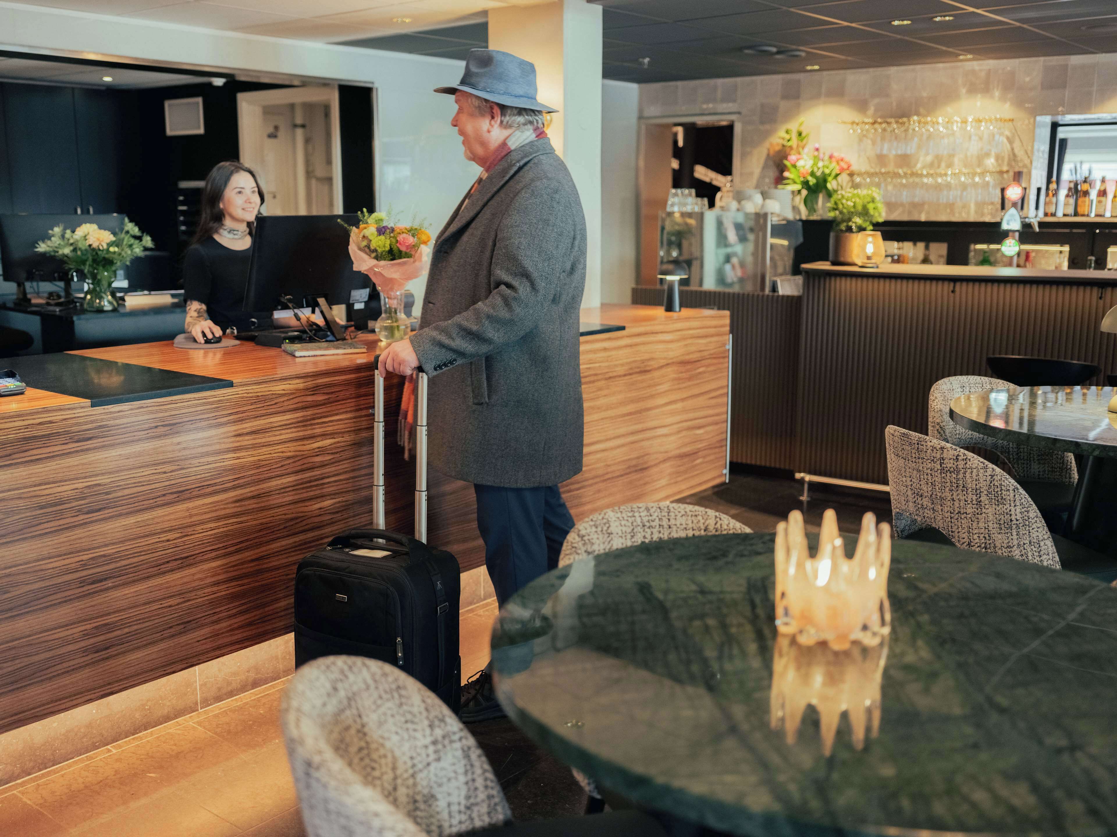 Zona de recepción de un hotel con un mostrador de madera, un bar con encimera de vidrio y un área de descanso con mesas y sillas redondas.