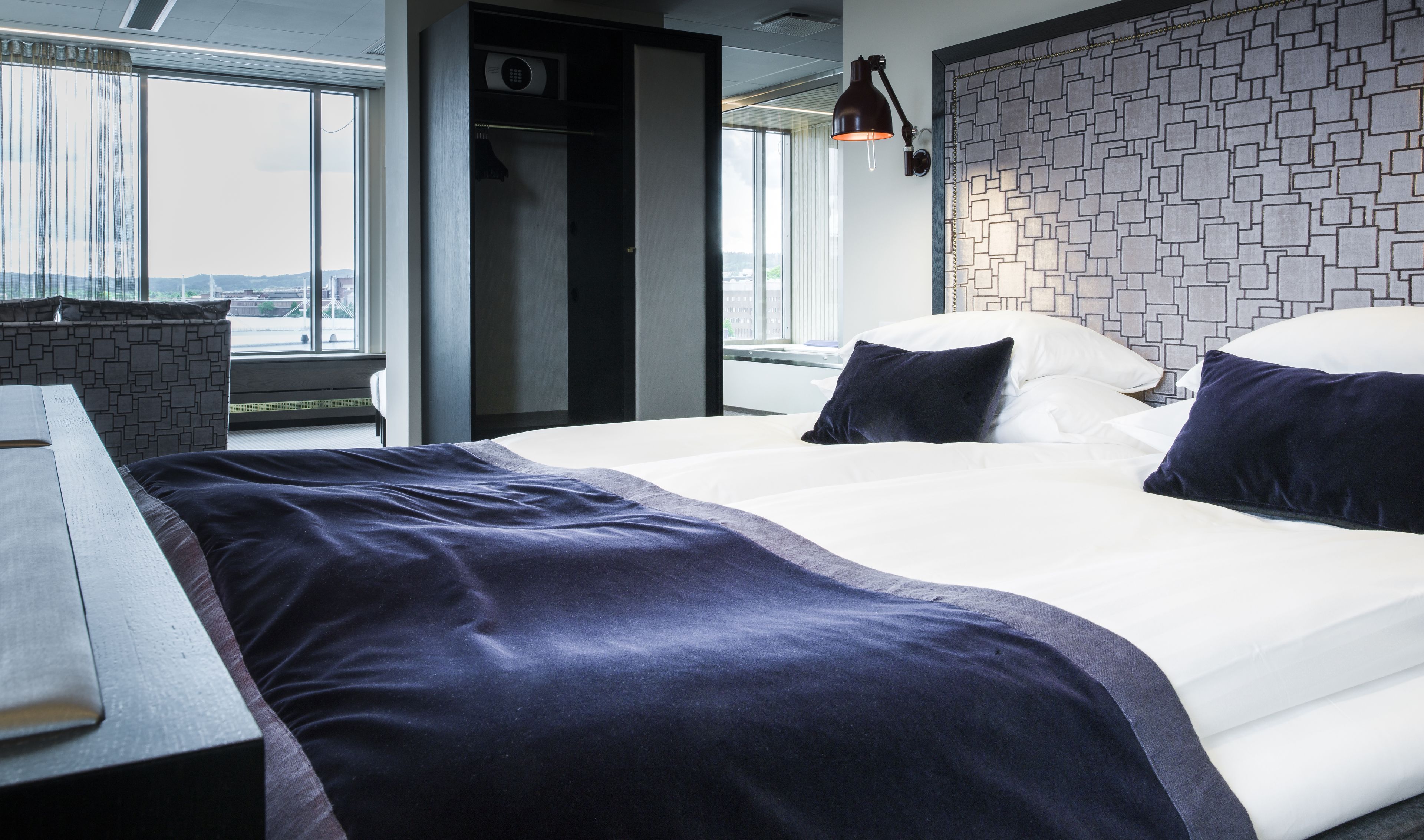 A modern hotel room with a large bed, a desk, and a view of the city through large windows.