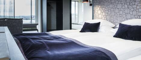 A modern hotel room with a large bed, a desk, and a view of the city through large windows.