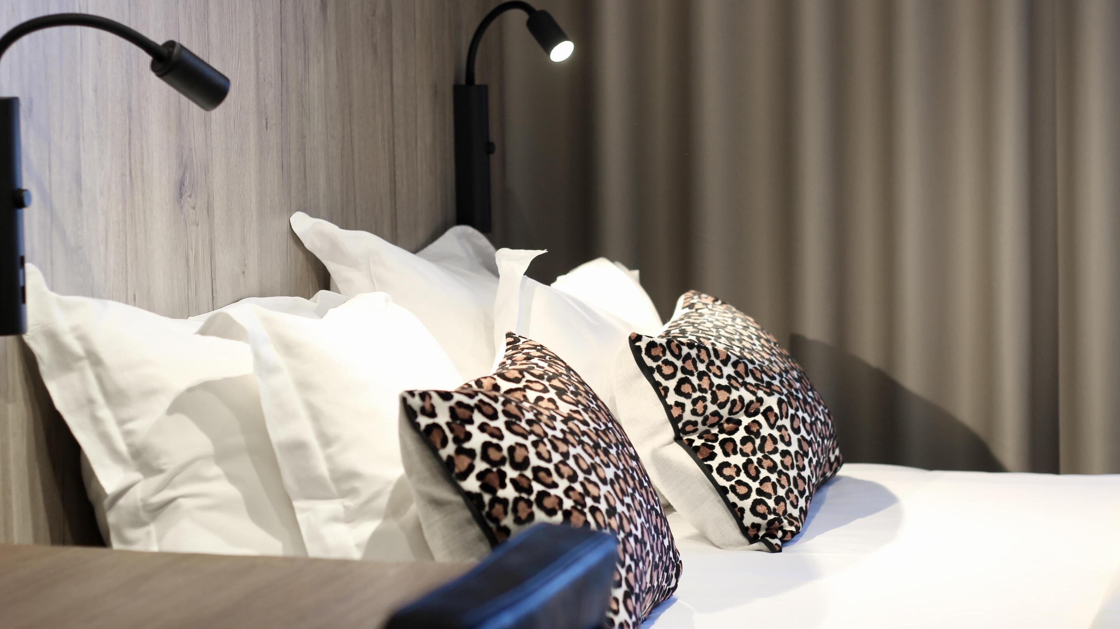Una cama bien hecha con almohadas blancas y de estampado de leopardo, un cabecero negro y una lámpara fijada a la pared.