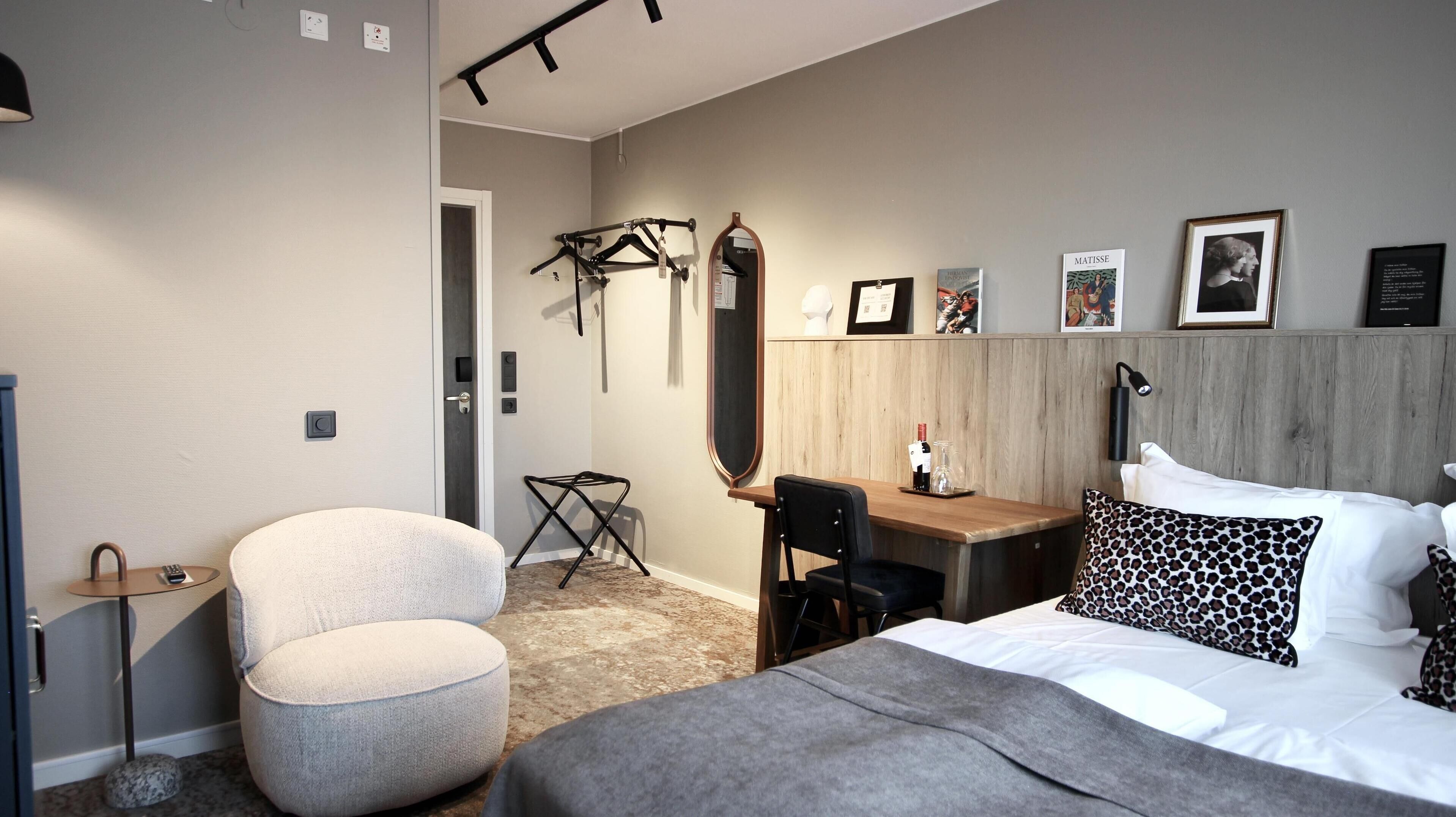 A modern hotel room with a bed, a desk, a chair, and a small table. There are wall-mounted lights, a mirror, and framed pictures.