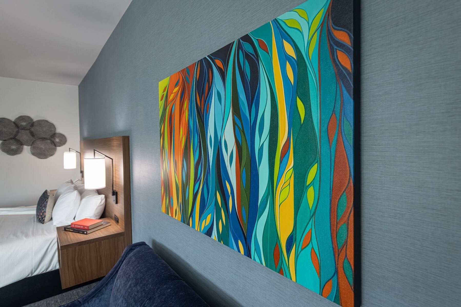 A hotel room with a large abstract painting on the wall, a bed with white linens, a bedside table with a lamp, and a blue sofa.