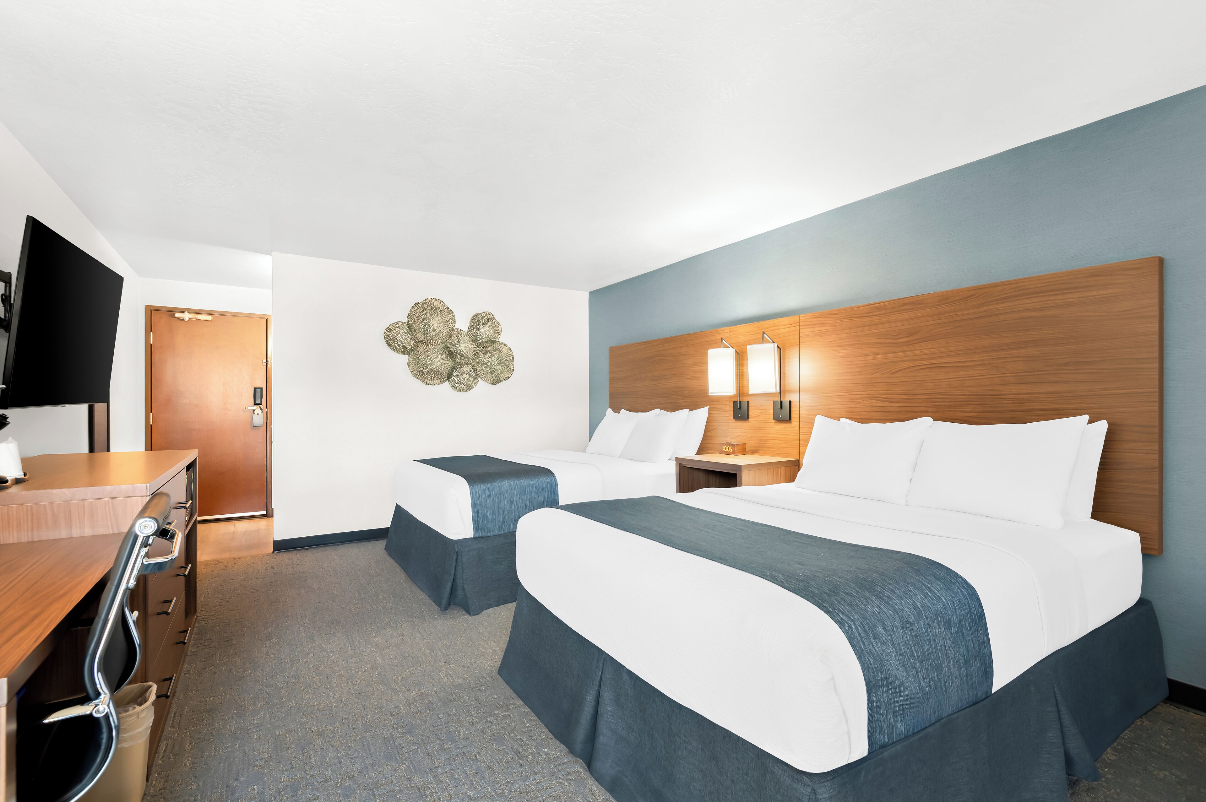 A hotel room with two beds, a wooden headboard, a flat-screen TV, a desk, and a wall-mounted light fixture.